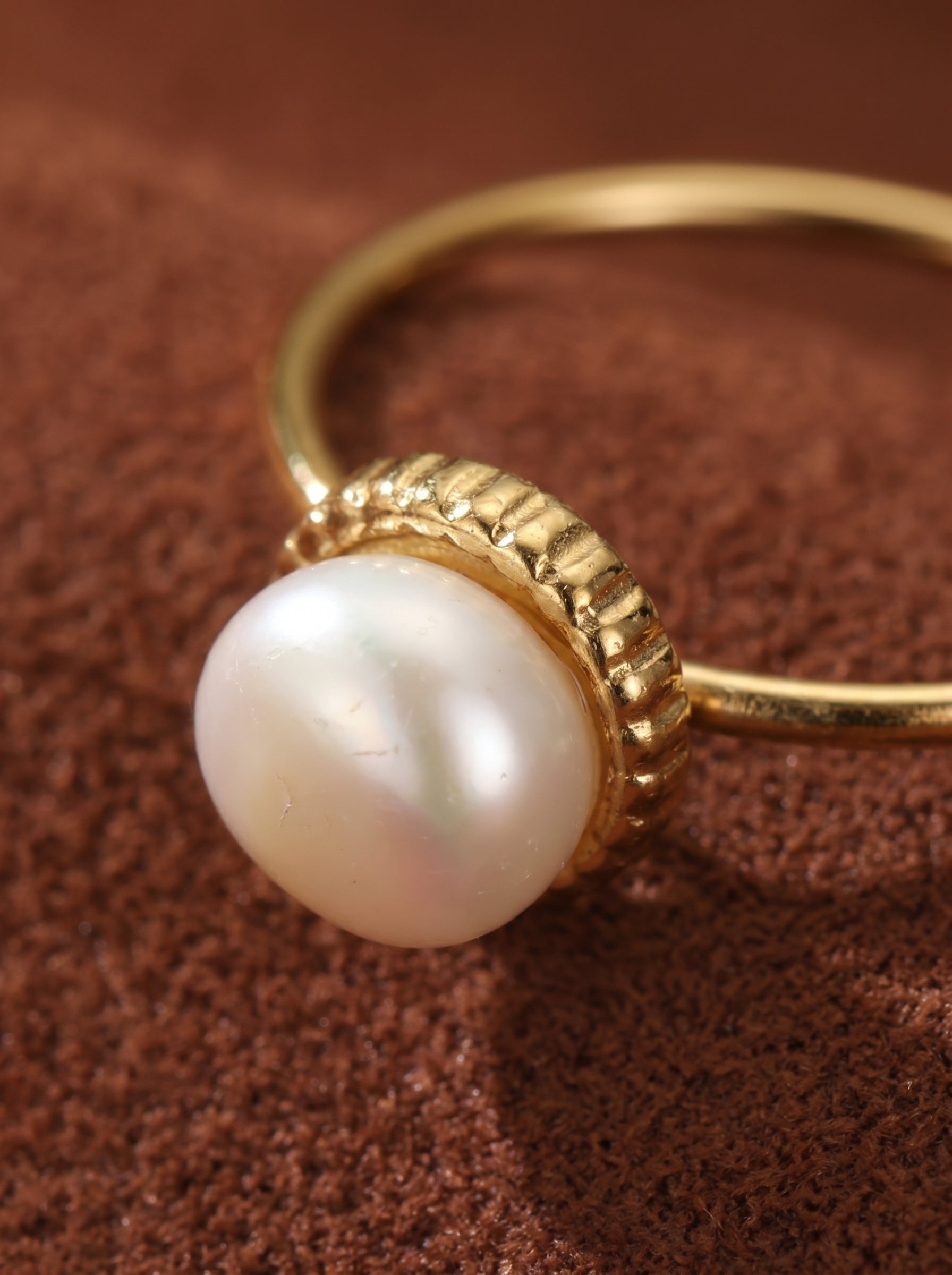 A close-up of a gold cuff ring with an open end, featuring a single, lustrous white freshwater button pearl set in a textured gold bezel. The ring rests on a textured brown surface. @pearl
