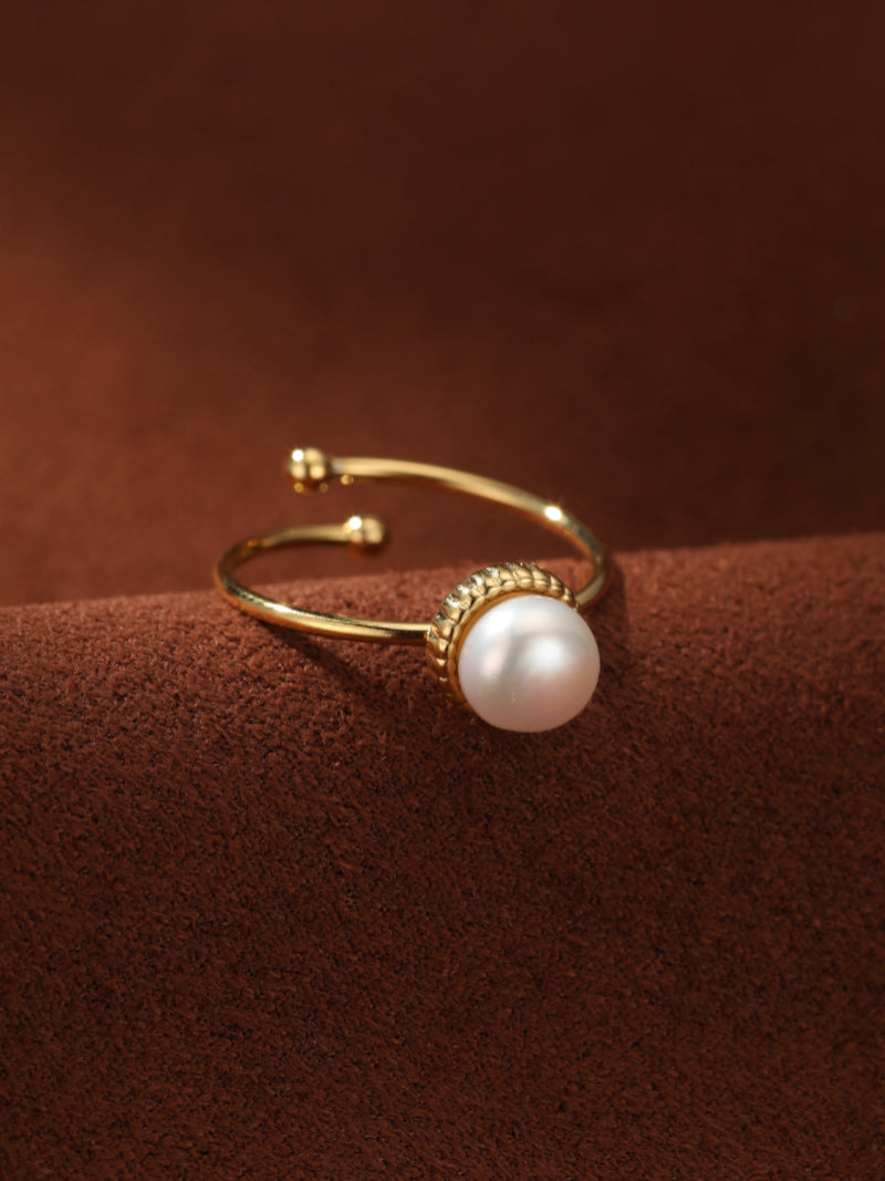 A close-up of a gold-toned open cuff ring featuring a single luminous 8 mm Freshwater White Button Pearl. The textured band of the ring is visible, and it rests on a rich brown textured surface.