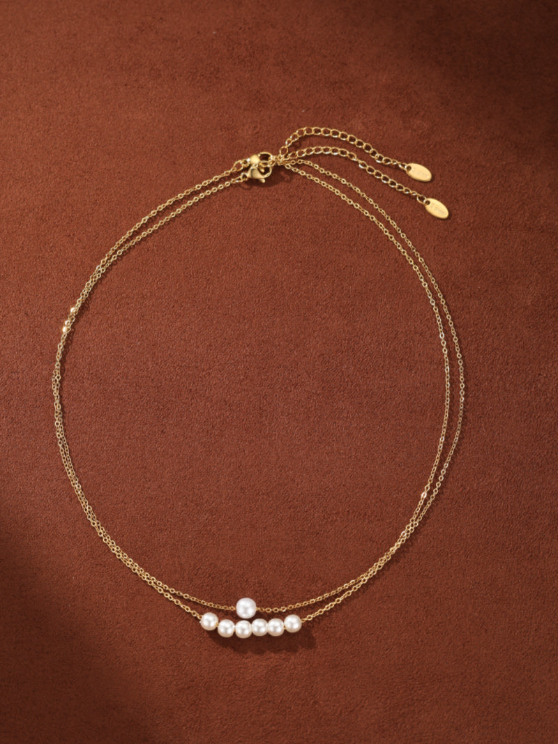 A delicate, gold-toned, layered chain necklace featuring a row of small, white, round imitation pearls on the lower chain, set against a brown background.