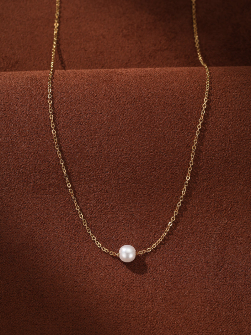 A delicate gold chain necklace with a single, lustrous 6mm round white imitation pearl centered on the chain. The necklace is displayed against a textured brown background.