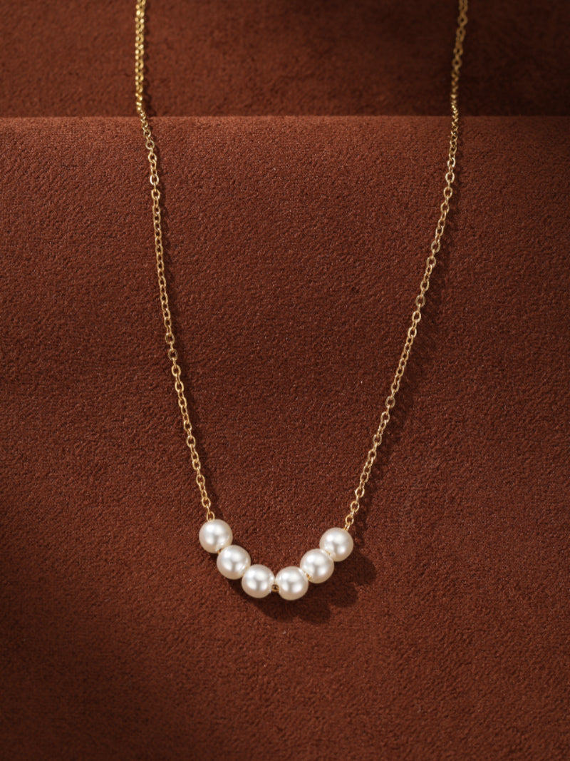 Delicate gold chain necklace with seven small, round, white imitation pearls arranged in a gentle curve. Product is shown against a textured brown background.