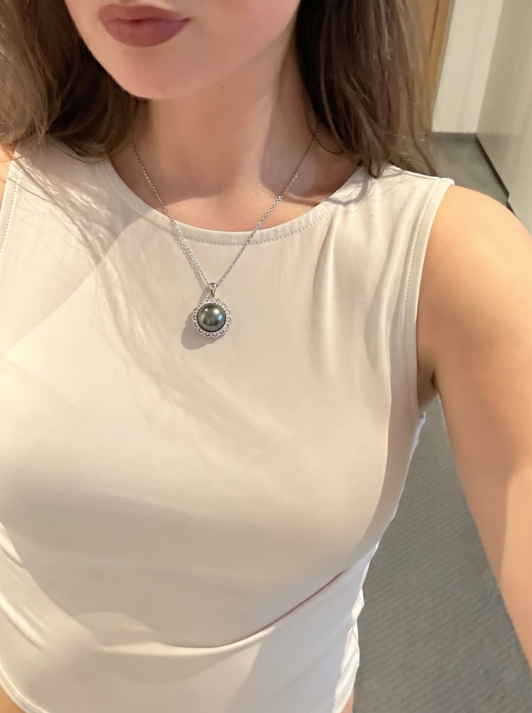A woman wearing a white sleeveless top, showcasing the Luminary Tahitian Mabe Pearl Sun Pendant Necklace. The necklace features a large, dark Tahitian Mabe pearl surrounded by a halo of sparkling accents, resembling a sunburst. @lifestyle