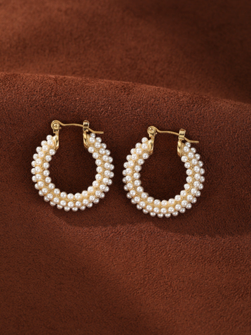 Gold-toned chunky hoop earrings densely adorned with multiple rows of lustrous white imitation pearls on a brown textured background.