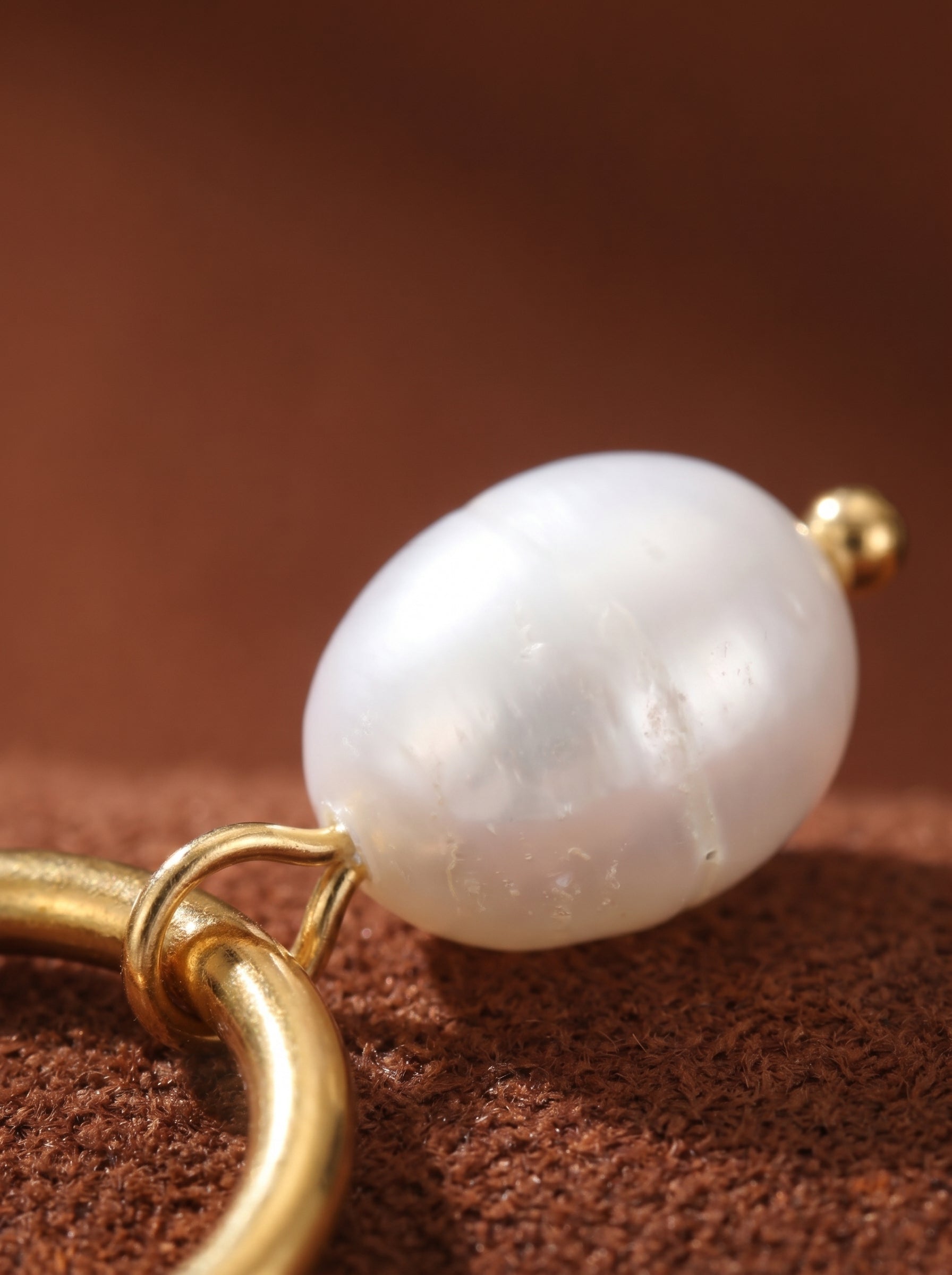 Close-up of a single Luminous Flow earring with a polished gold-tone hoop and a dangling white freshwater pearl. The pearl has a lustrous sheen and is accented by a small gold bead. The earring is set against a warm brown textured background. @pearl