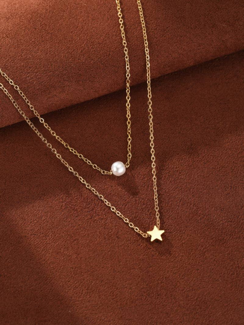 Collier superposé ton or avec une perle ronde d'eau douce sur une chaîne et une breloque étoile sur l'autre, présenté sur fond texturé marron.