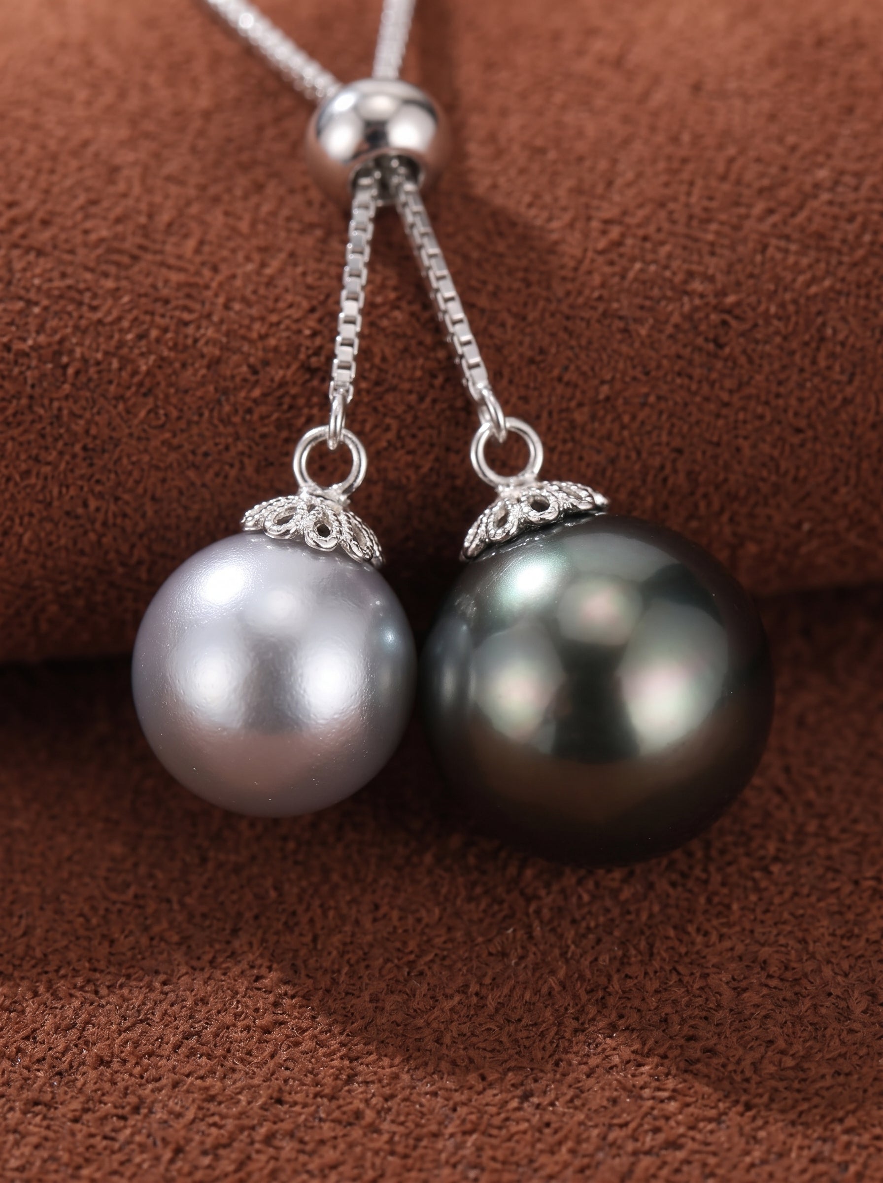 Close-up of a Y-drop pendant necklace featuring a lustrous black Tahitian pearl and a bright white Akoya pearl, suspended on a silver chain against a textured brown background. @pearl