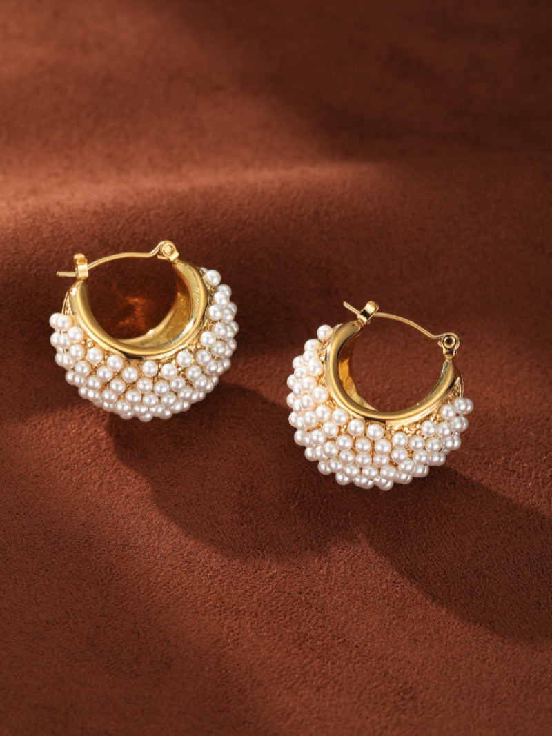 Lush Freshwater Pearl Hoop Earrings featuring a gold hoop encrusted with numerous small white freshwater pearls. The earrings have a contemporary, architectural design and are shown against a brown textured background.