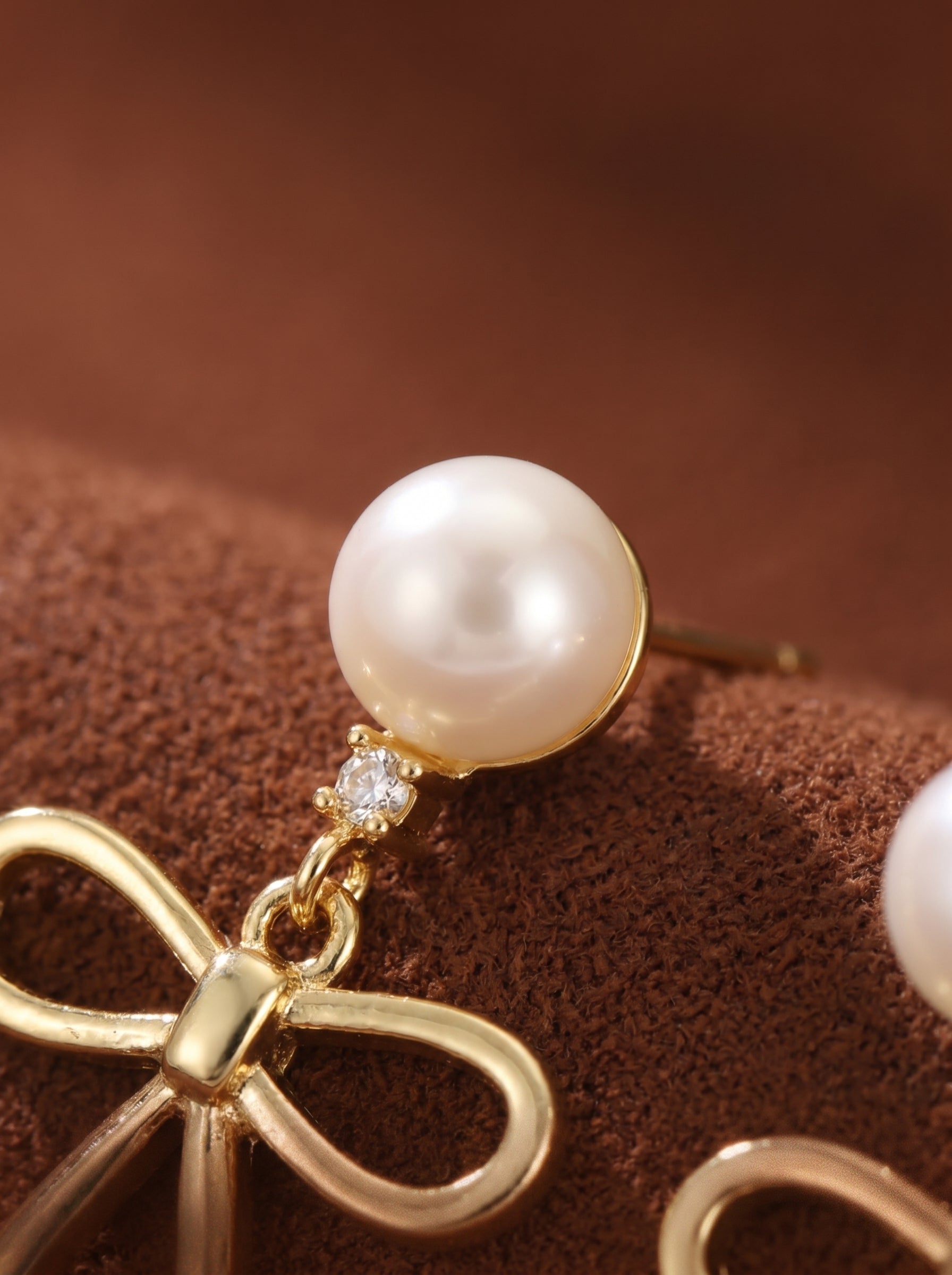 Gold-toned drop earrings featuring a lustrous white freshwater pearl stud with a small clear stone, connected by a delicate chain to a polished bow charm. @pearl