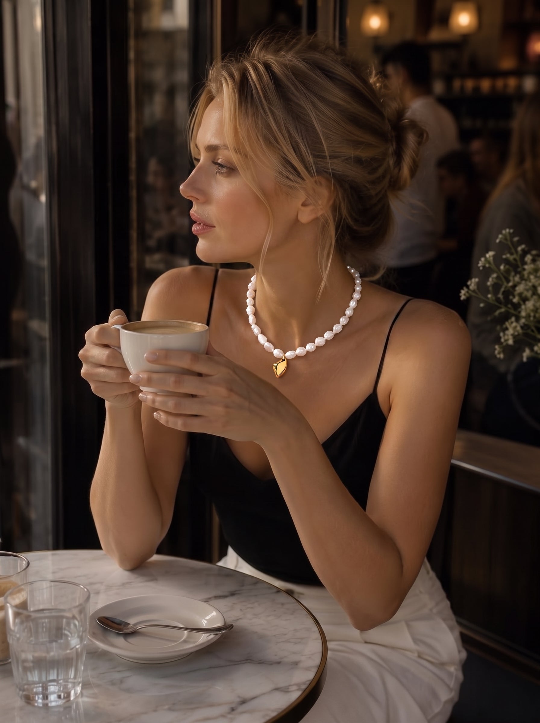 Une femme portant une caraco noir et un pantalon blanc est assise à une table en marbre dans un café, tenant une tasse de café. Elle est parée du collier pendentif perlé Lyrical en perle d'eau douce, présentant un fil de perles blanches irrégulières et un pendentif goutte abstrait lisse ton or. @lifestyle