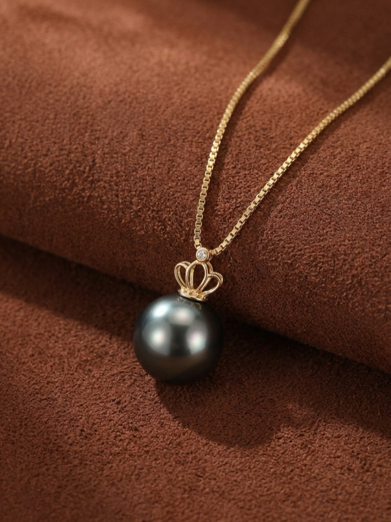 A close-up studio shot of the Majesty Tahitian Pearl Crown Pendant Necklace, featuring a single, lustrous 9mm round Tahitian pearl topped with a delicate gold crown motif, suspended from a fine gold box chain. A small diamond accent is visible above the pearl. The necklace rests on a textured brown background.
