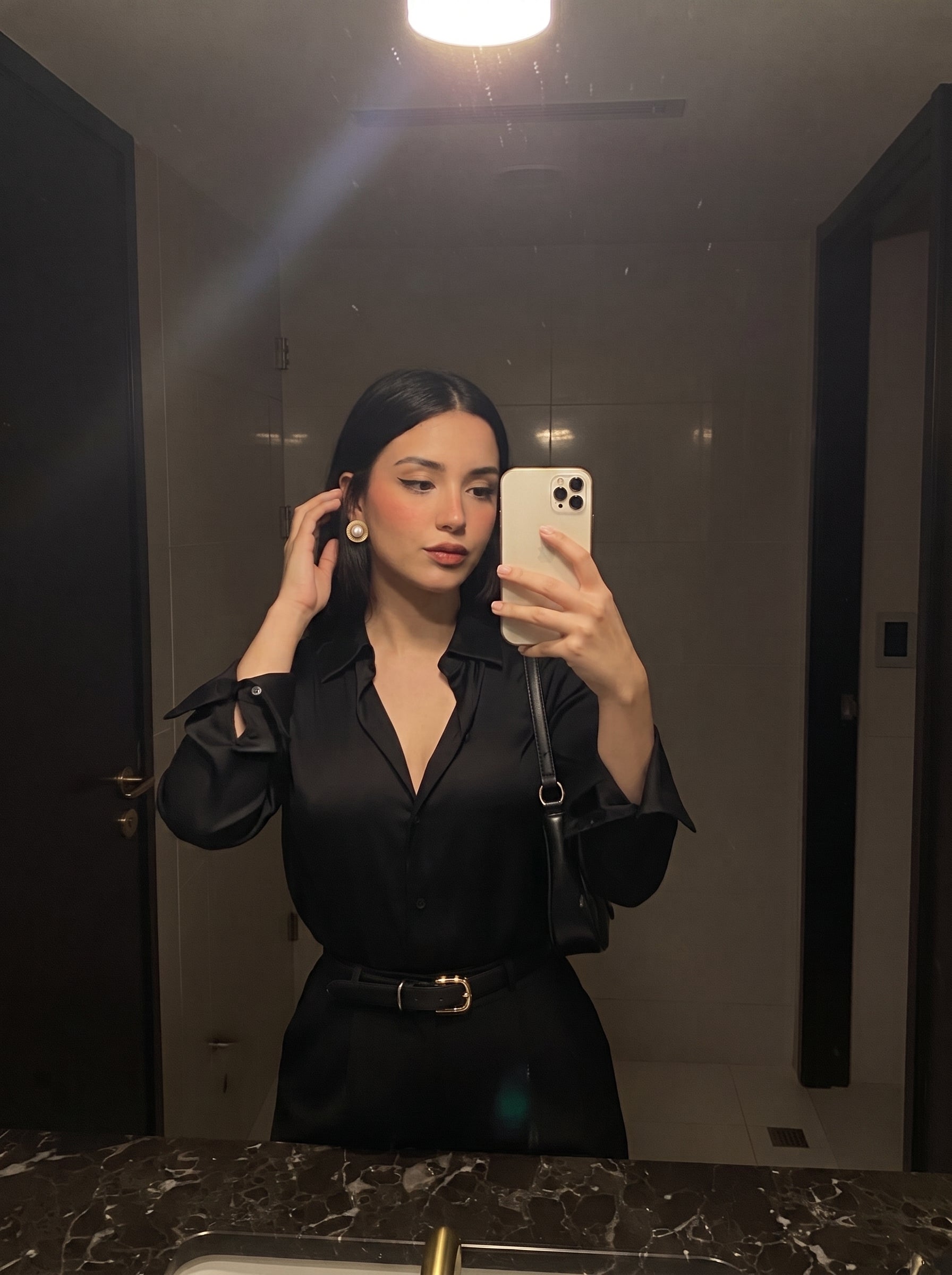 A woman in a black satin shirt takes a mirror selfie, showcasing her Meridian Shine Freshwater Pearl Sunburst Stud Earrings. The earrings feature a white pearl surrounded by a textured gold sunburst frame. @lifestyle