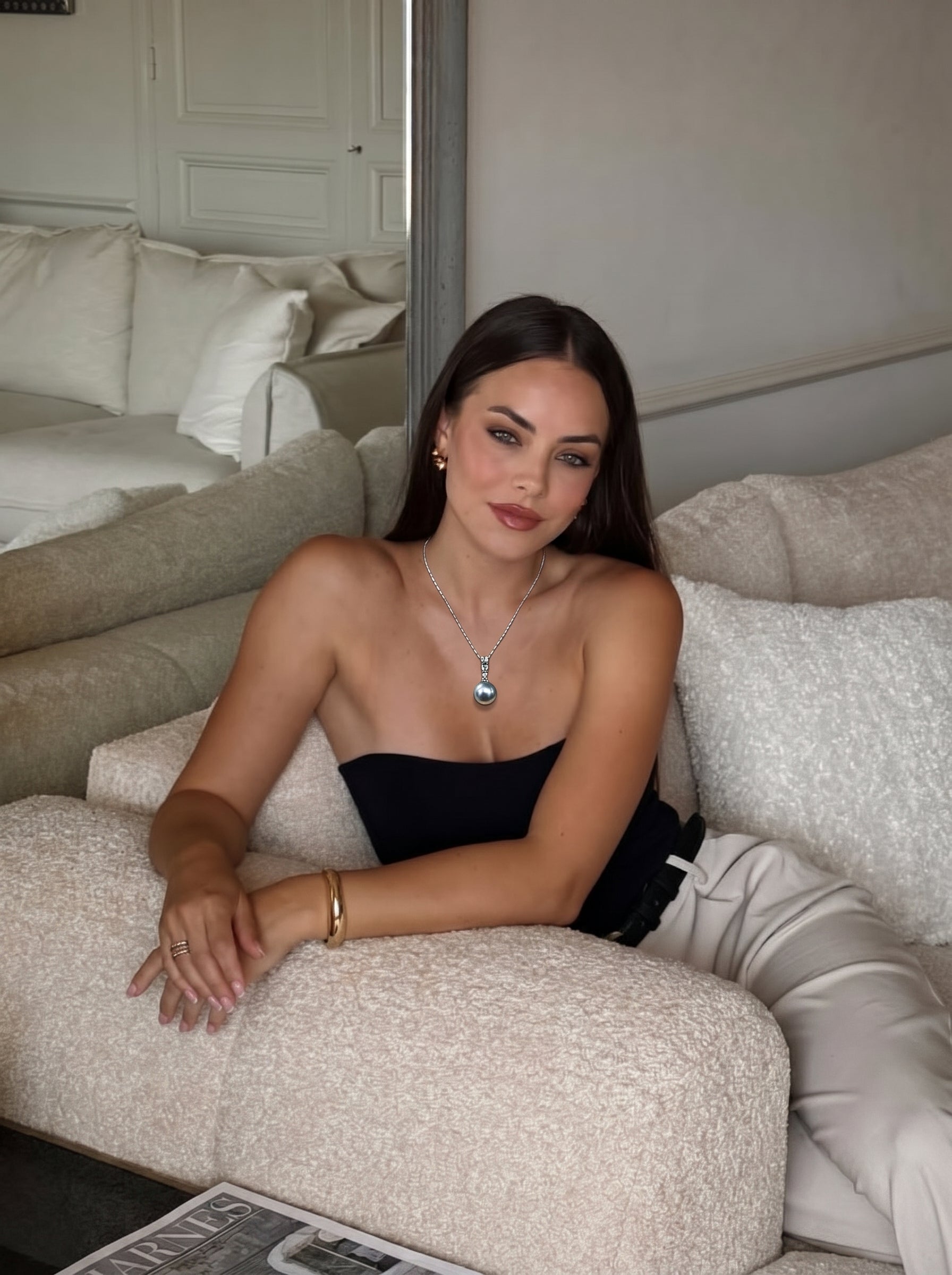 Une femme portant le collier pendentif à chaîne Moonbeam en perle Akoya avec une perle Akoya argentée ronde de 9 mm et une bélière scintillante, assise sur un canapé crème. @lifestyle