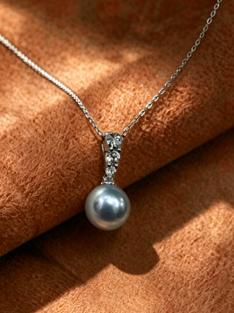 Moonbeam Akoya Pearl Drop Chain Necklace featuring a single, luminous 9mm round silver Akoya pearl suspended from a delicate silver chain with a sparkling diamond-accented bail on a textured brown background.