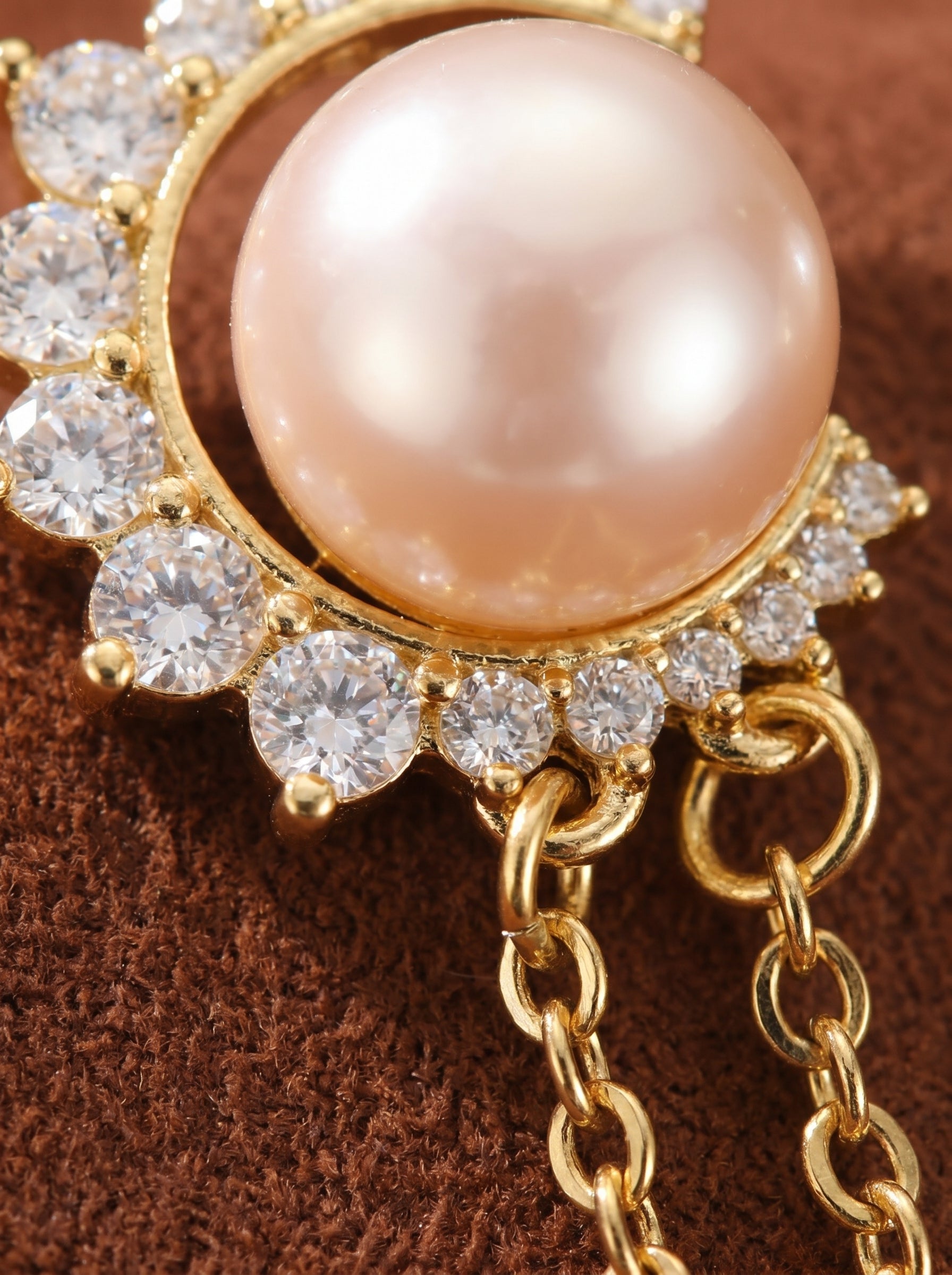 Close-up of a gold dangle earring featuring a large, light pink freshwater pearl set within a crescent moon shape adorned with clear sparkling cubic zirconia stones, with delicate gold chains extending downwards. @pearl