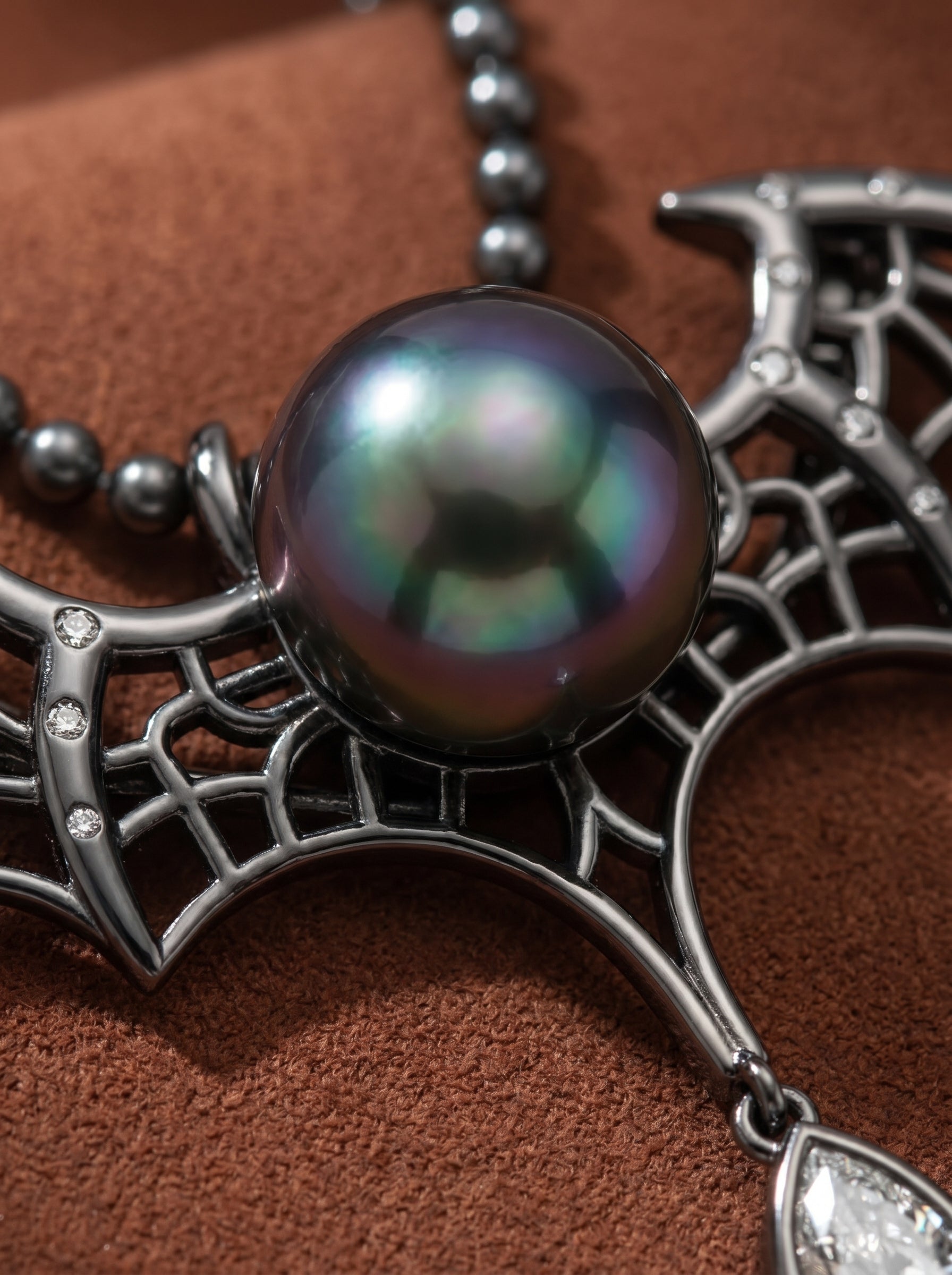 A close-up shot of the Nocturne necklace, showcasing a large, iridescent Tahitian pearl as the centerpiece, nestled within an intricate black metal bat wing pendant adorned with small crystals. The necklace features a beaded chain and a pear-shaped crystal drop. @pearl