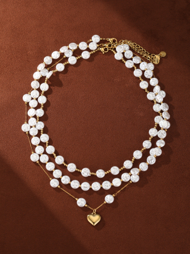 Layered necklace featuring three strands of white baroque freshwater pearls accented with small gold beads, with a delicate gold heart pendant on the lowest strand.