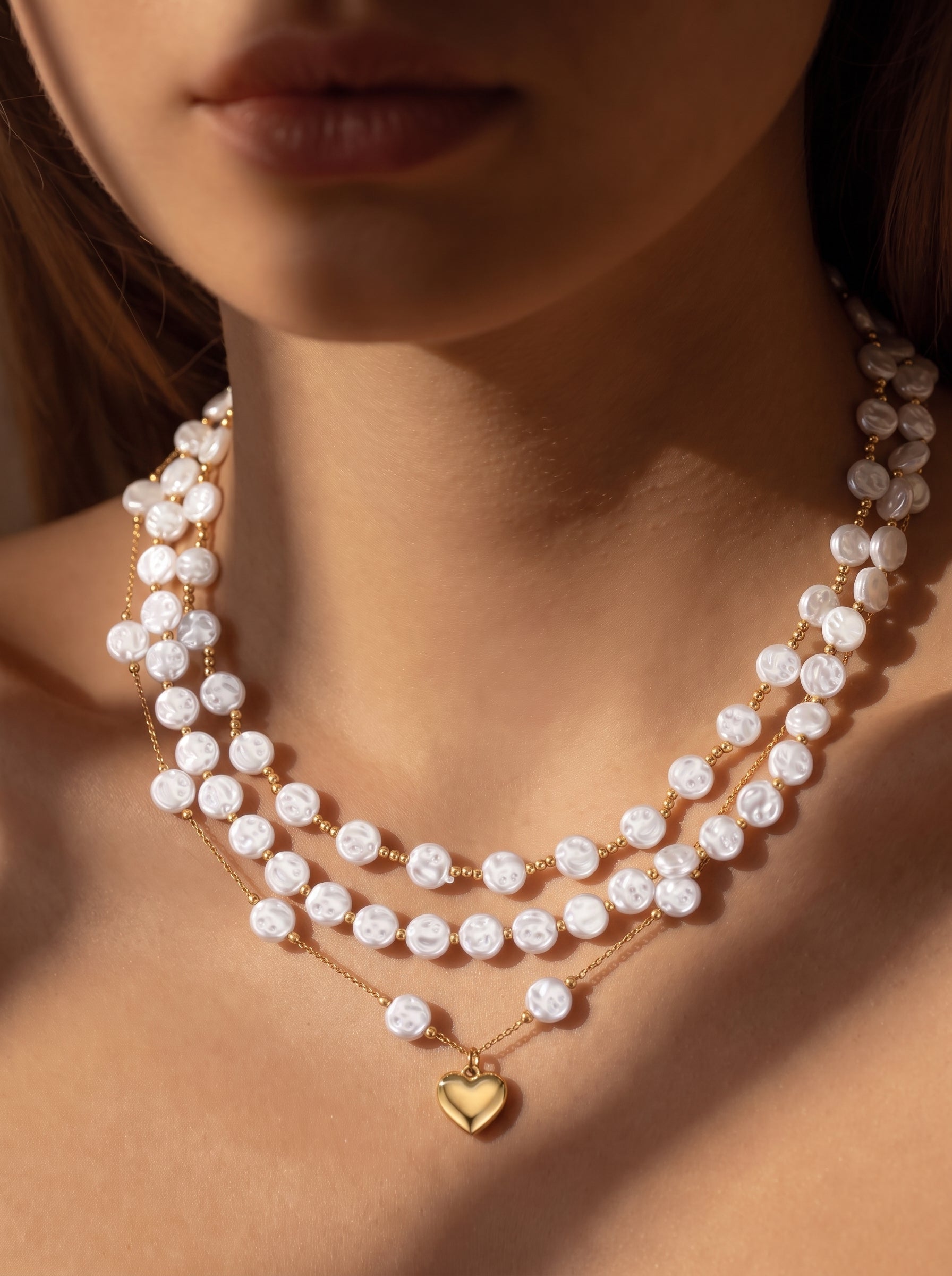 A layered necklace featuring three strands of white freshwater baroque pearls with gold bead accents and a gold heart pendant, worn by a model. @model