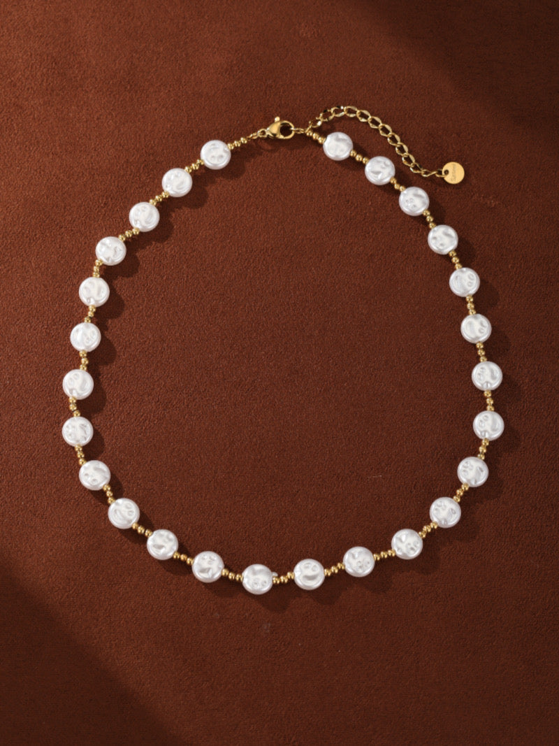 A gold-toned necklace featuring a single strand of alternating white baroque freshwater pearls and small gold beads, laid out on a brown textured surface.