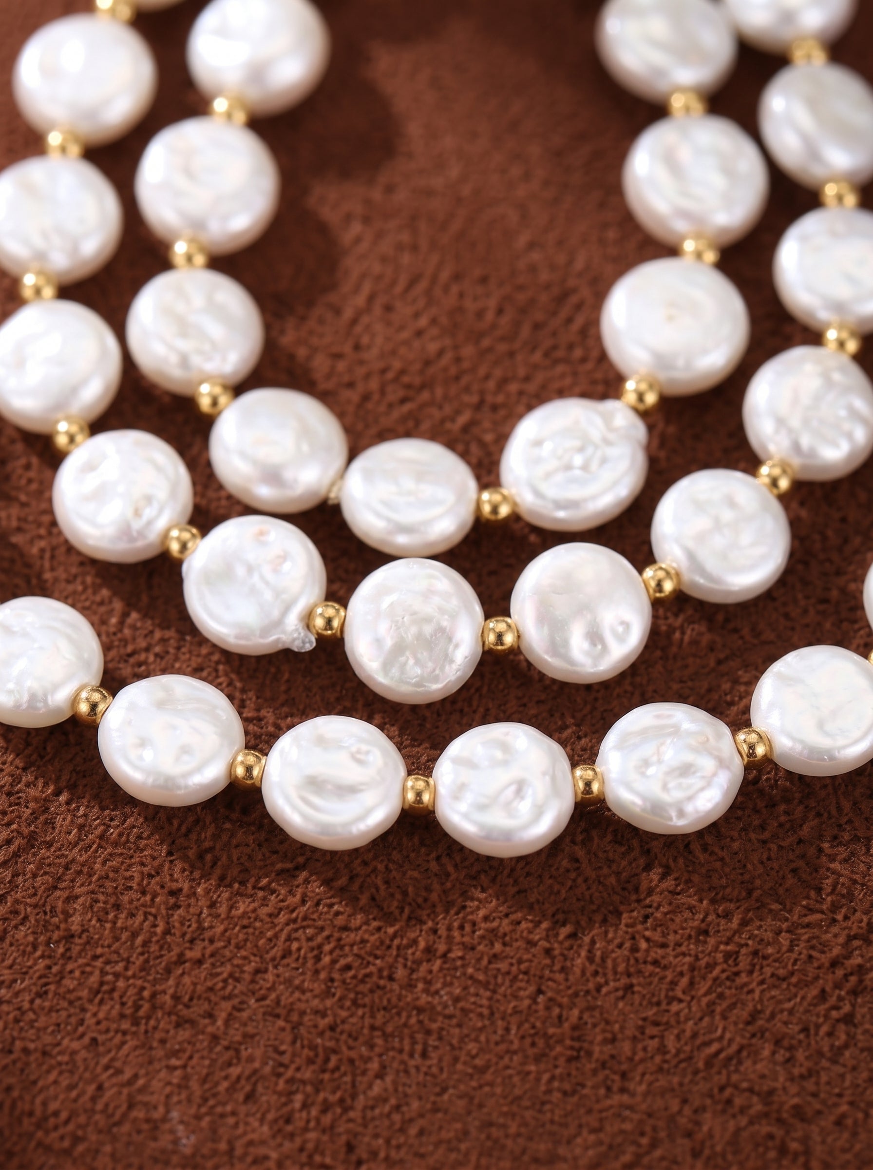 Close-up of a layered necklace featuring three strands of flat, white freshwater pearls with gold bead spacers, set against a brown textured background. @pearl