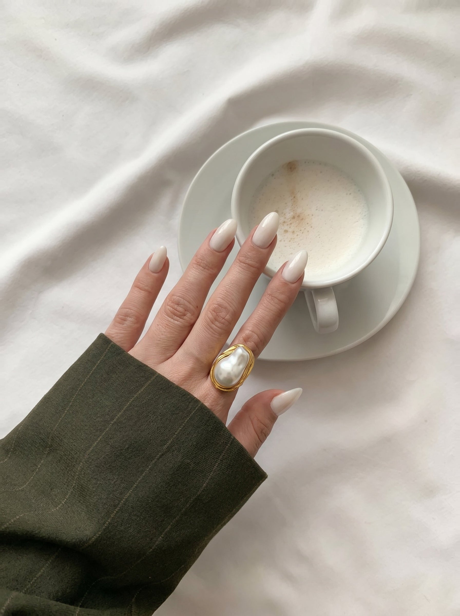 A gold-toned cuff ring with a large, irregular white baroque freshwater pearl sits on a woman's finger, next to a cup of coffee on a white surface. @lifestyle #lifestyle