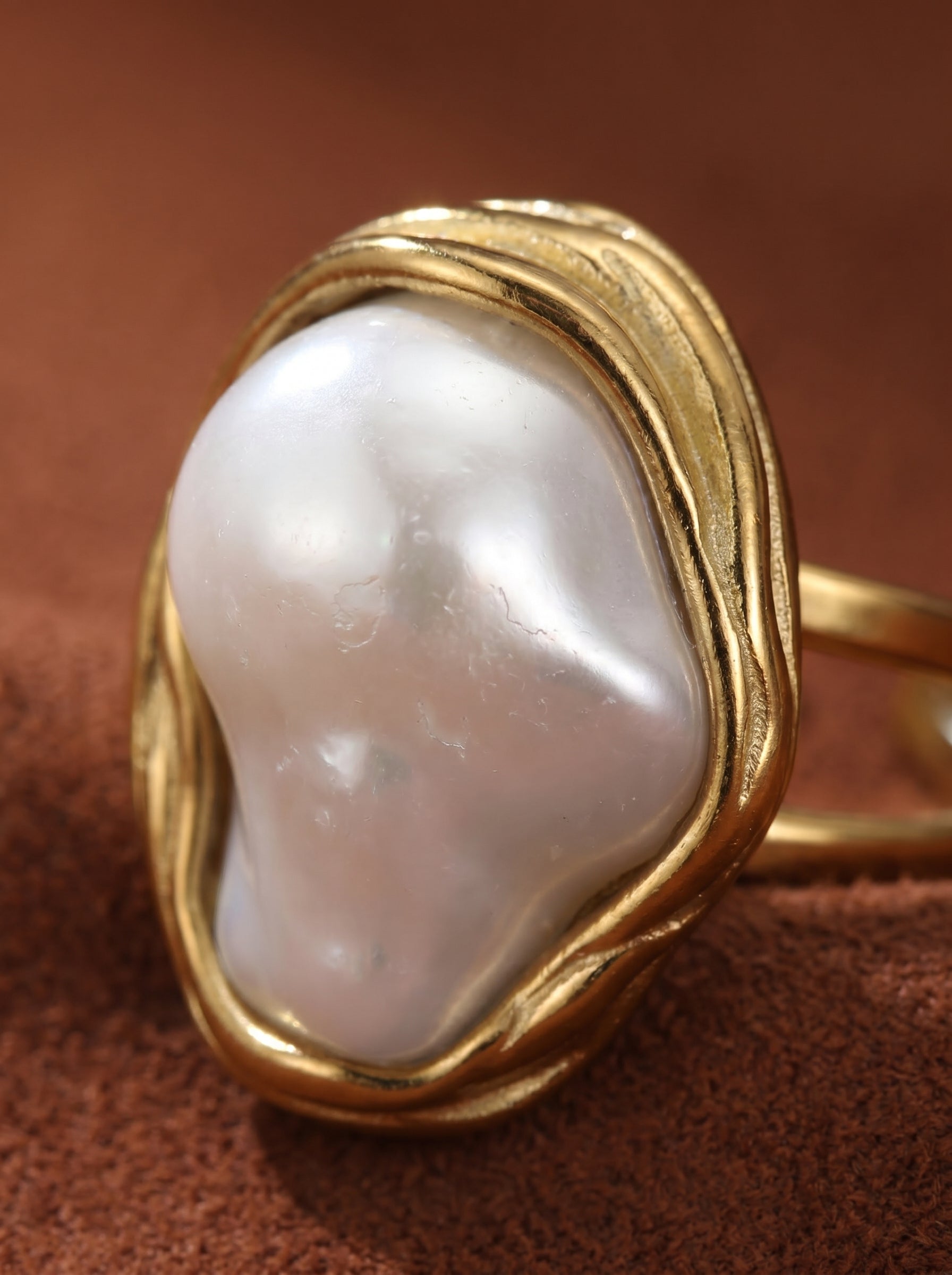 Close-up of a white baroque freshwater pearl set in an organic gold-toned cuff ring on a textured brown background. @pearl