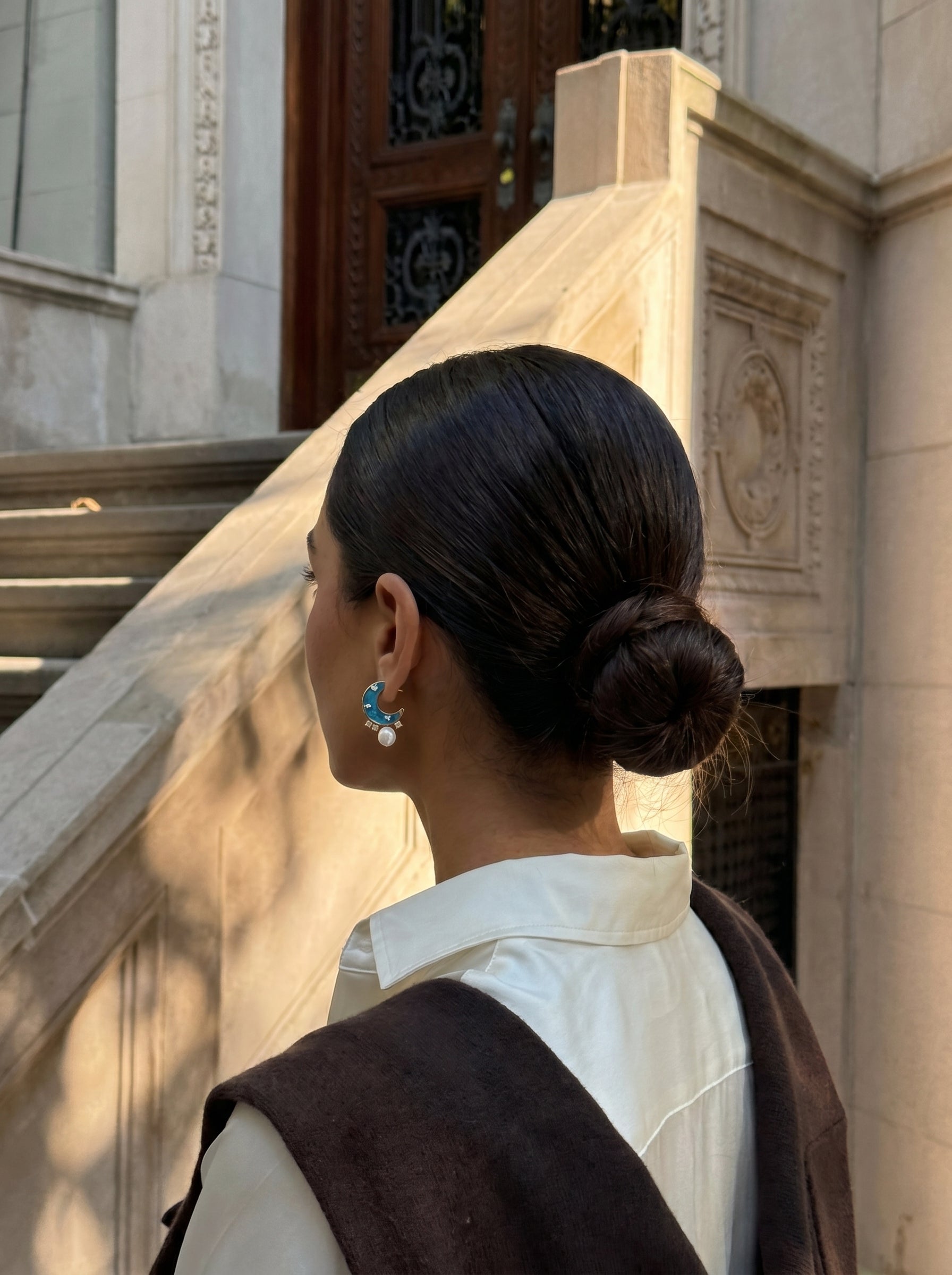 Gros plan d'une femme portant les boucles d'oreilles puces lune et étoile Orbit en perle d'eau douce avec des détails en coquille d'ormeau irisée, à l'extérieur près d'escaliers en pierre. @lifestyle