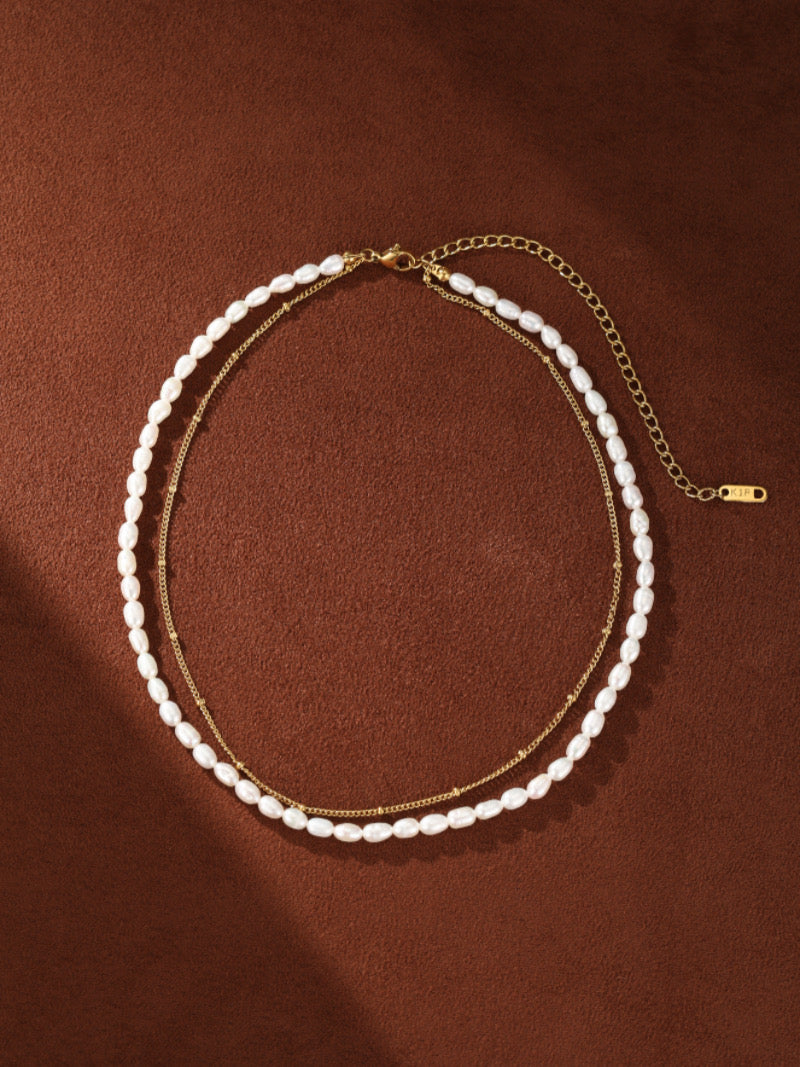 Un délicat collier superposé présentant un fil de petites perles ovales blanches d'eau douce et une fine chaîne ton or, posé à plat sur fond texturé marron.