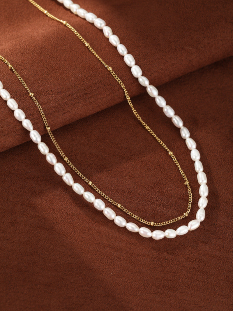 Deux colliers superposés, l'un avec des perles graines blanches d'eau douce et l'autre une délicate chaîne ton or avec de petites perles, présentés sur fond texturé marron.