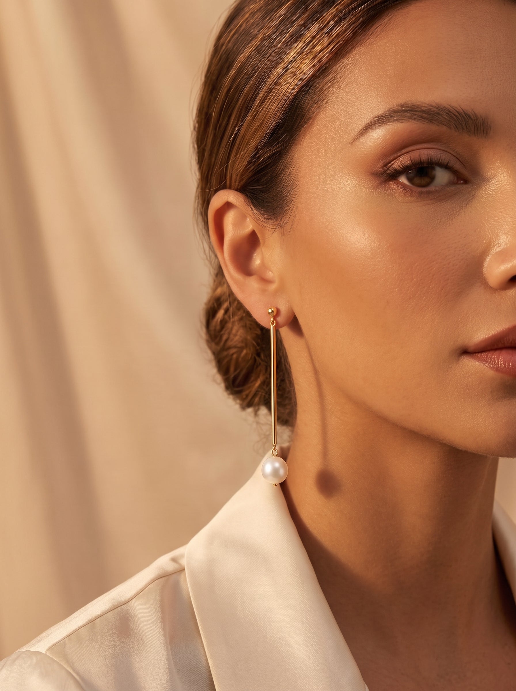 Gros plan de l'oreille d'une femme portant une boucle d'oreille goutte ton or avec une grande perle blanche d'eau douce pendant d'une fine barre. Le fond est un rideau doux rose pêche avec de subtiles ombres, suggérant un cadre sophistiqué. @lifestyle