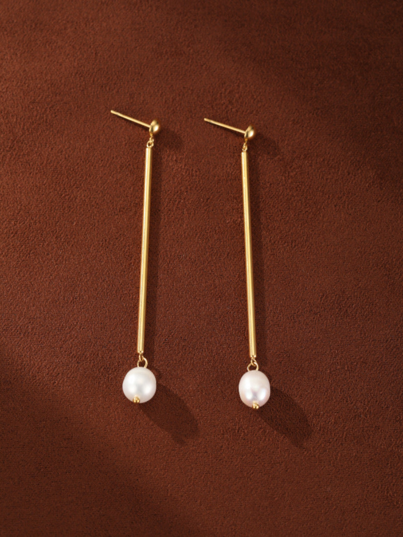 Boucles d'oreilles goutte barre ton or présentant de lustrées perles blanches d'eau douce sur fond marron