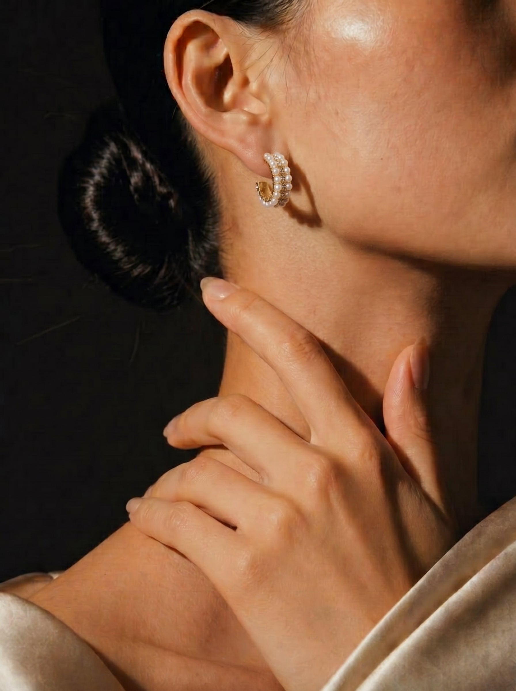 Close-up of a model wearing gold hoop earrings adorned with multiple rows of small, lustrous freshwater pearls and sparkling crystals. The earrings are positioned on her ear, with her hand gently touching her neck. @model