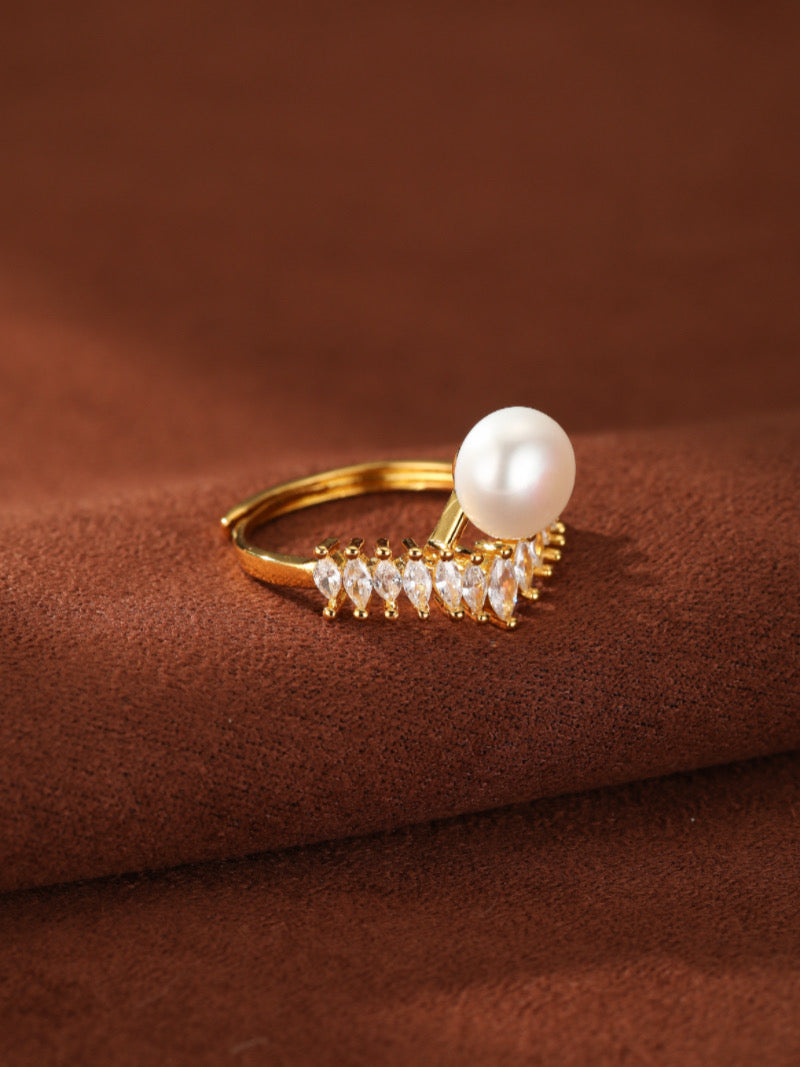 Gold cuff ring featuring a white freshwater pearl and a V-shaped band adorned with marquise-cut clear stones, on a brown textured background.