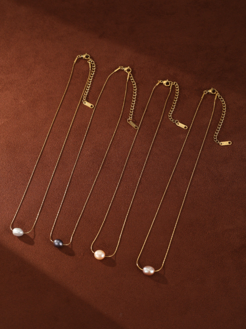 Quatre colliers pendentif goutte unique Poise en perle d'eau douce en perles blanches, noires, roses, et rose pâle sur de délicates chaînes dorées, présentés sur fond marron.
