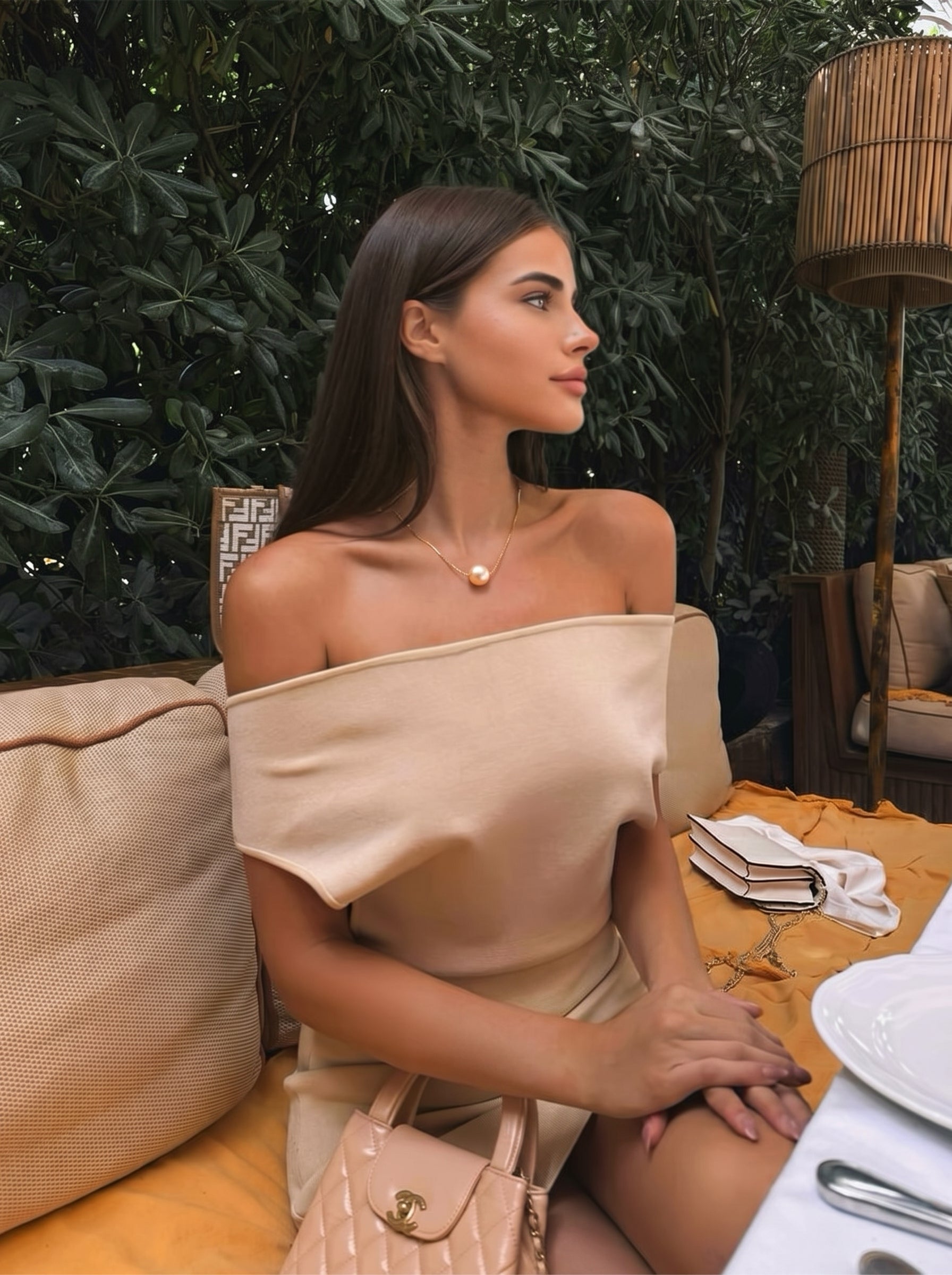 Femme portant le collier pendentif goutte unique Poise en perle d'eau douce en perle blanche, assise en extérieur à une table avec une verdure luxuriante en arrière-plan. @lifestyle