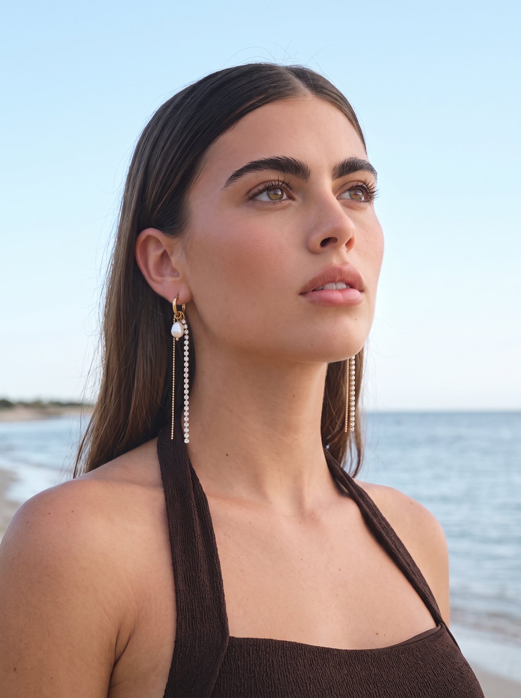 A woman wearing Prism Freshwater Pearl Dangle Earrings at the beach, with the ocean in the background. The earrings feature a gold hoop, a white pearl, and a dangling line of crystals. @lifestyle