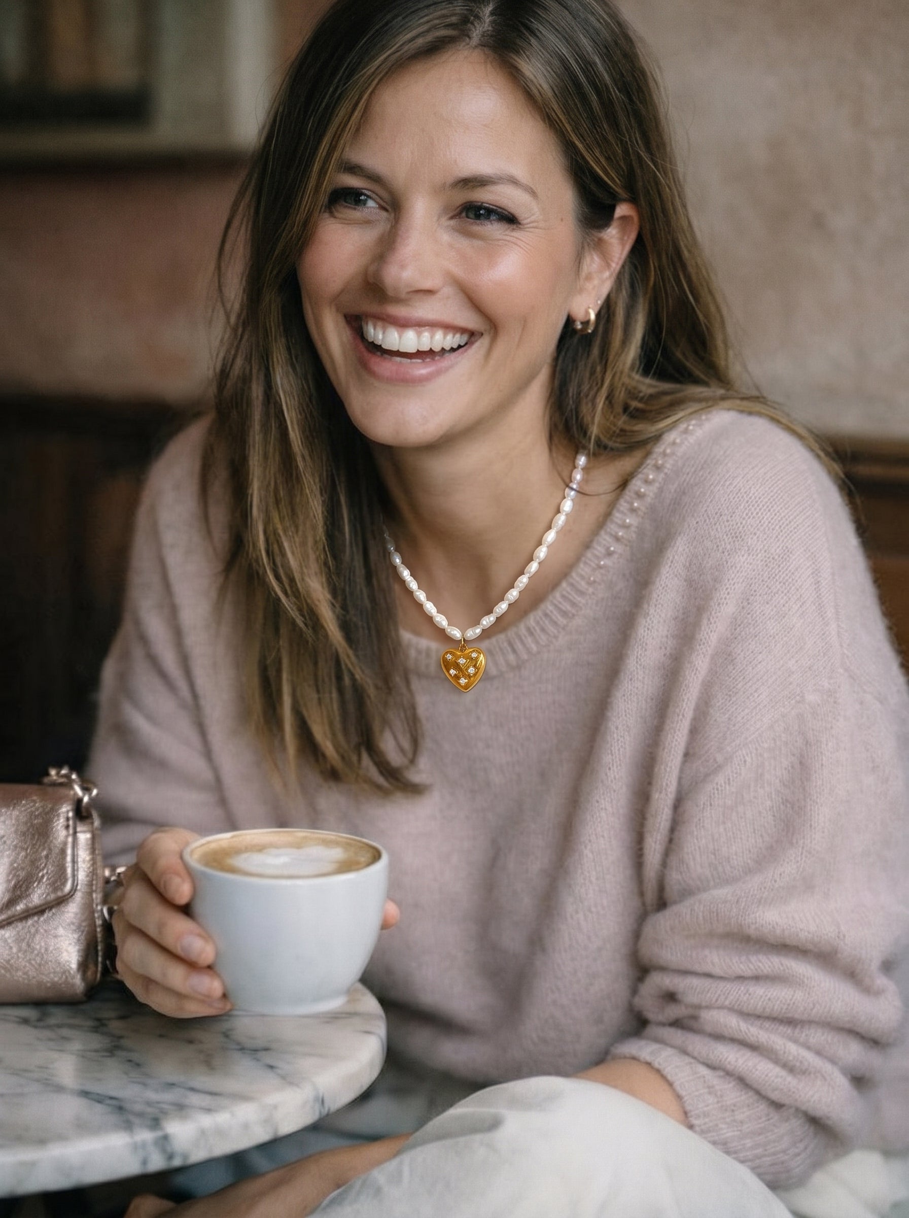 Smiling woman wearing a white freshwater pearl choker necklace with a gold heart pendant, holding a coffee cup at a cafe. @lifestyle #lifestyle