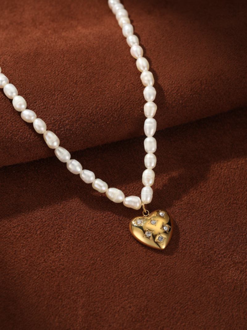 Close-up of a necklace featuring a strand of oval freshwater pearls leading to a gold-tone heart pendant adorned with small sparkling stones. @pearl
