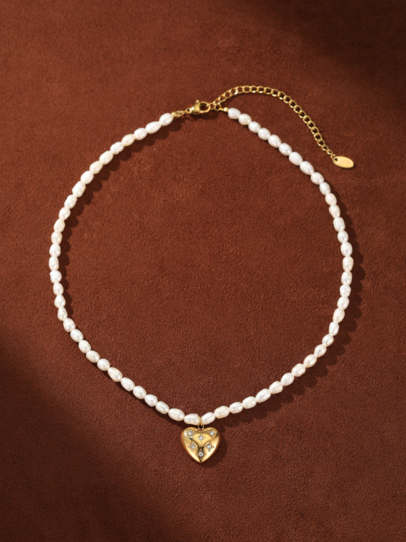 A choker necklace featuring a strand of small, oval freshwater pearls and a gold heart pendant adorned with sparkling accent stones. The necklace has a gold clasp and extender chain. @pearl