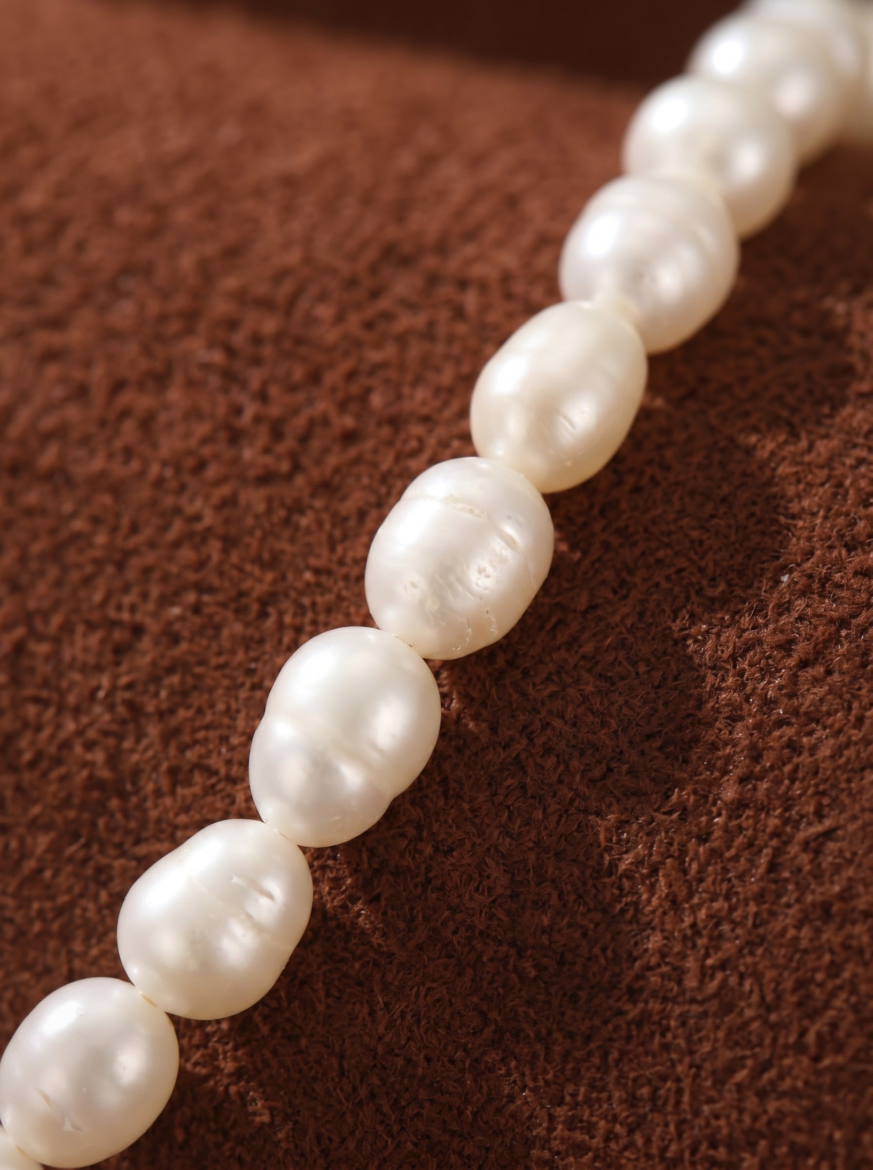 A string of lustrous, irregular white freshwater pearls with a subtle sheen, laid diagonally on a textured brown fabric background. The pearls are approximately 4mm and showcase their natural, organic shape. @pearl