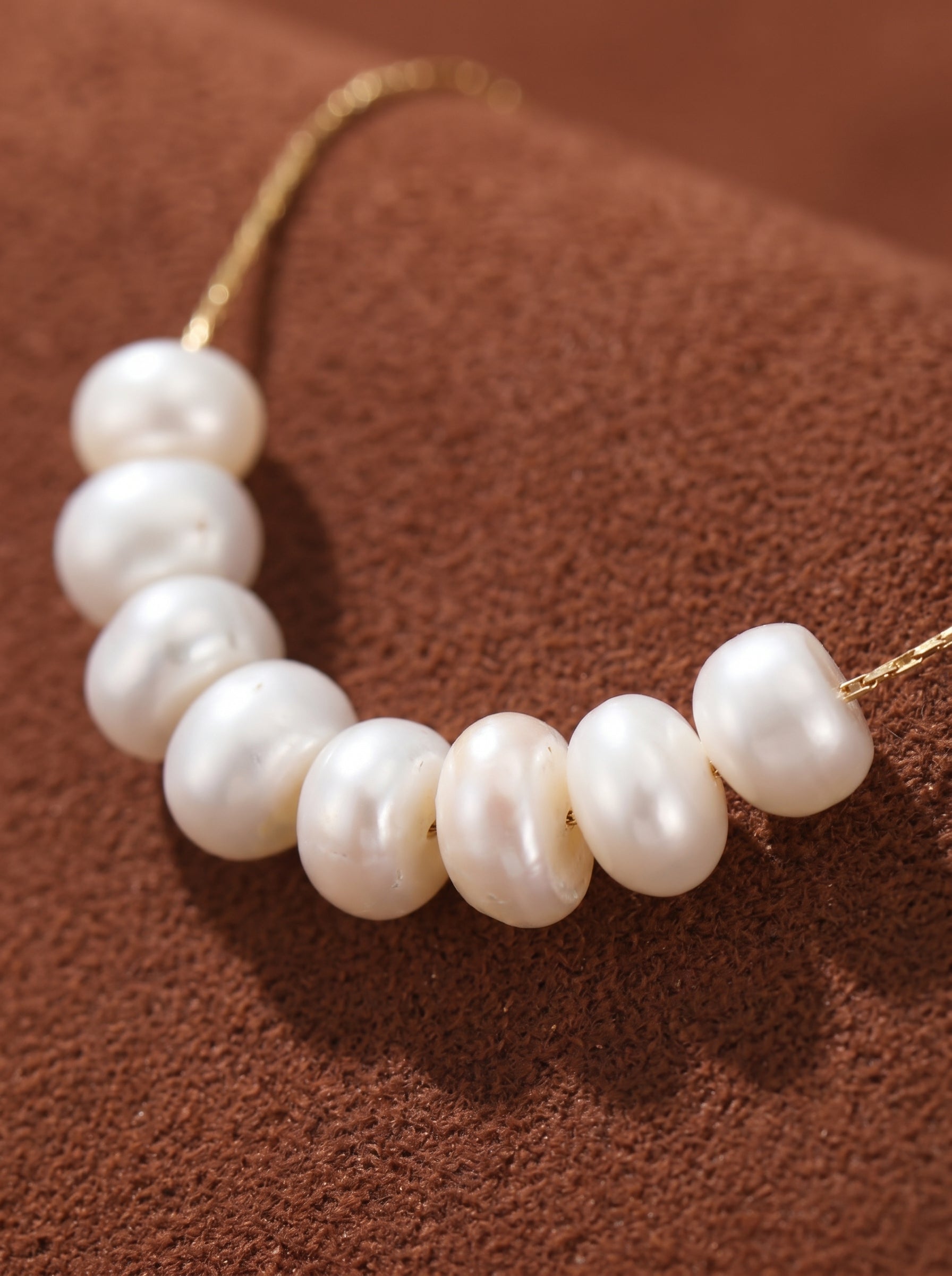 Delicate gold chain necklace with seven lustrous 4mm white button freshwater pearls appearing to float along the chain, against a brown textured background. @pearl