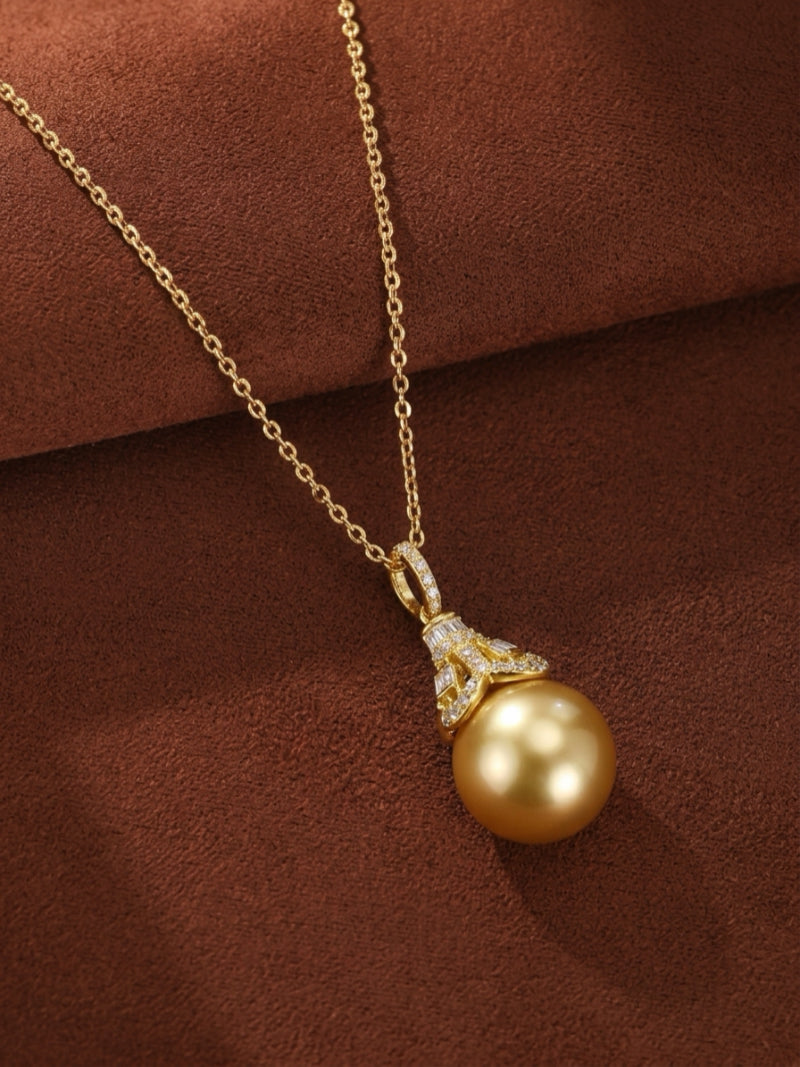 A close-up studio shot of the Regal Glow pendant necklace featuring a large, lustrous 14mm round golden South Sea pearl suspended from a delicate yellow gold chain. The pearl is capped with an ornate, diamond-encrusted bail showcasing both round and baguette-cut diamonds.
