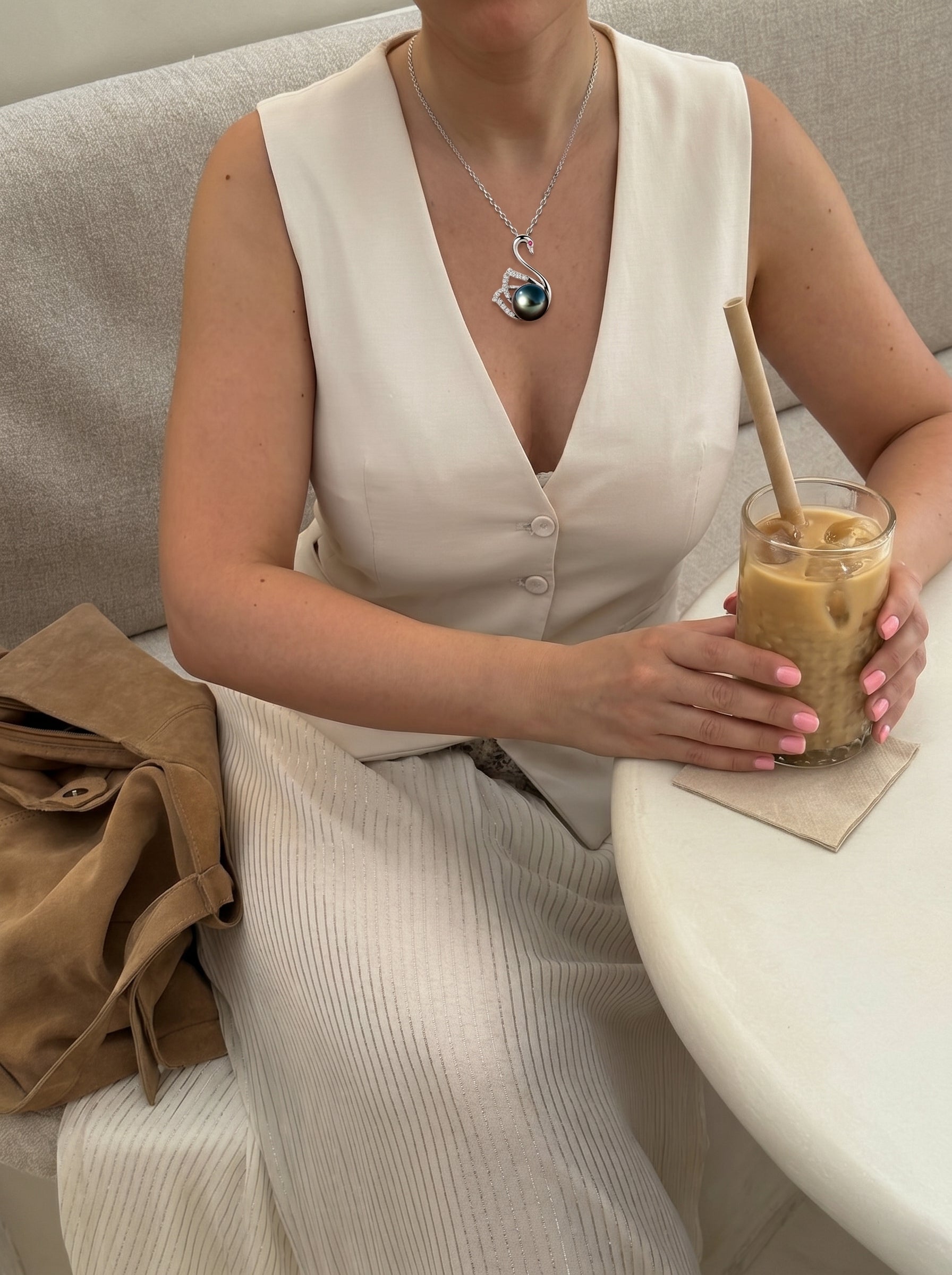 Une femme en gilet et jupe blancs tient un café glacé tout en portant le collier pendentif cygne Reverie en perle de Tahiti, présentant un pendentif cygne en argent sterling avec des accents diamant berçant une perle noire. @lifestyle