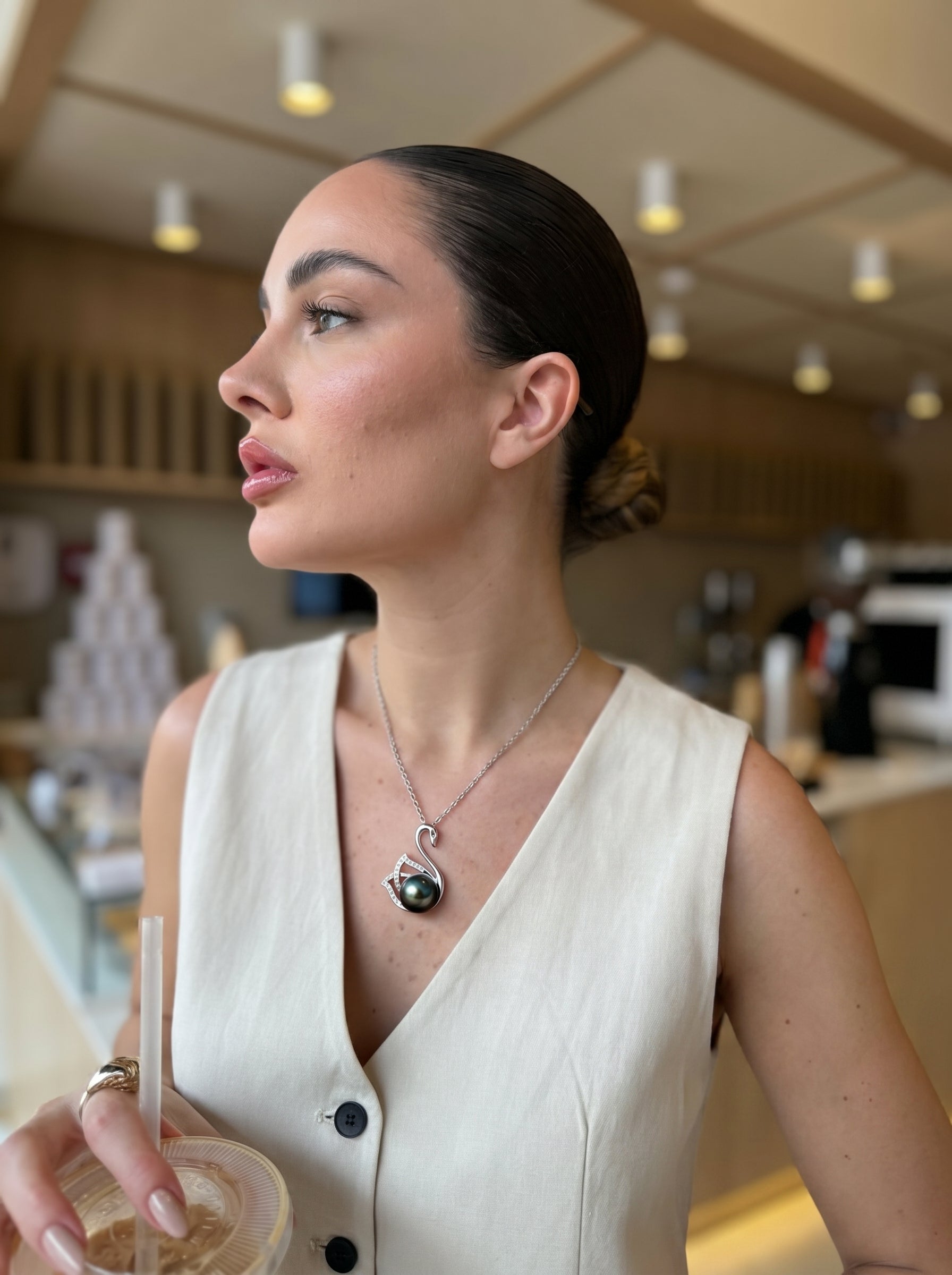 Une femme porte le collier pendentif cygne Reverie en perle de Tahiti, présentant un pendentif cygne en argent sterling avec des accents diamant retenant une lustrée perle noire de Tahiti. Elle est vêtue d'un gilet blanc et tient une boisson dans un cadre de café. @lifestyle