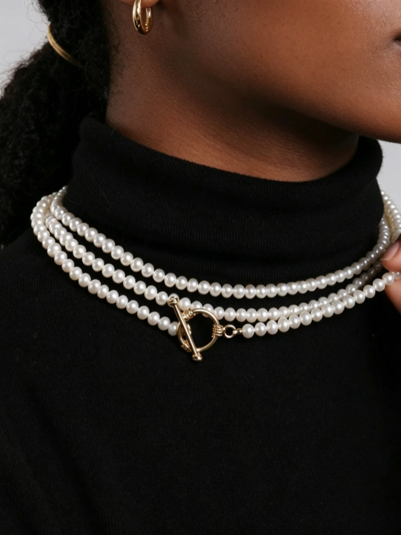 Close-up of a woman wearing a triple strand white freshwater pearl necklace with a gold toggle clasp over a black turtleneck. @model