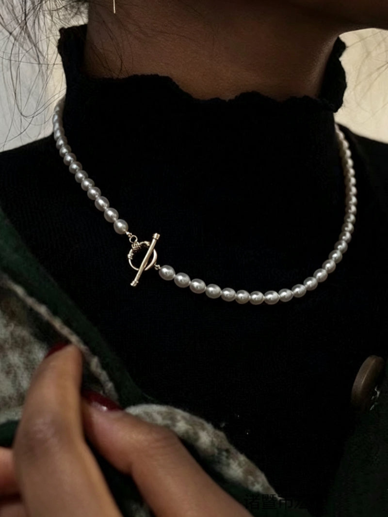 Close-up of a delicate, single strand of white freshwater pearls with a gold toggle clasp, worn over a black turtleneck. @model