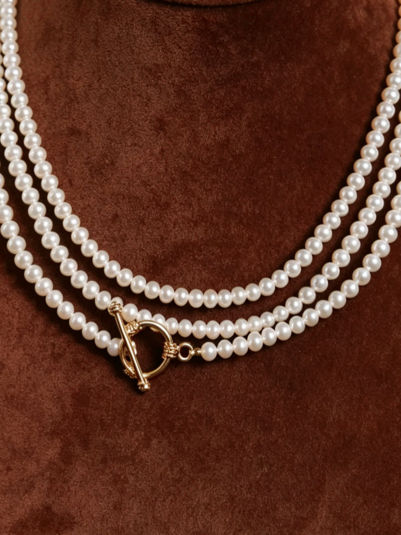 Close-up of a triple strand freshwater pearl necklace with a gold toggle clasp against a brown velvet background.