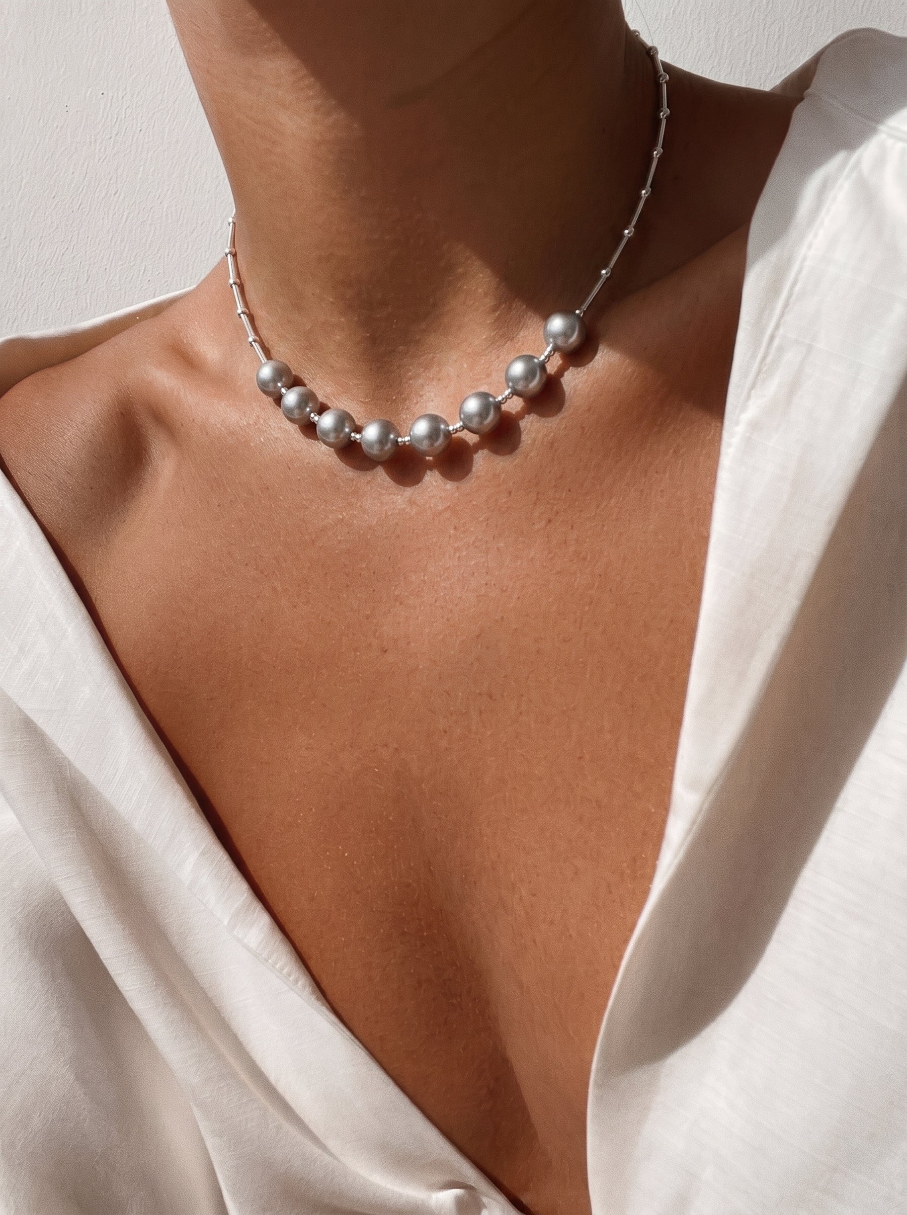 A close-up shot of a person wearing a white shirt and a delicate silver necklace with scattered Akoya pearls. The pearls are round and have a subtle sheen, spaced evenly along the chain. The necklace is positioned on the décolletage, highlighting its elegant and modern design. @model