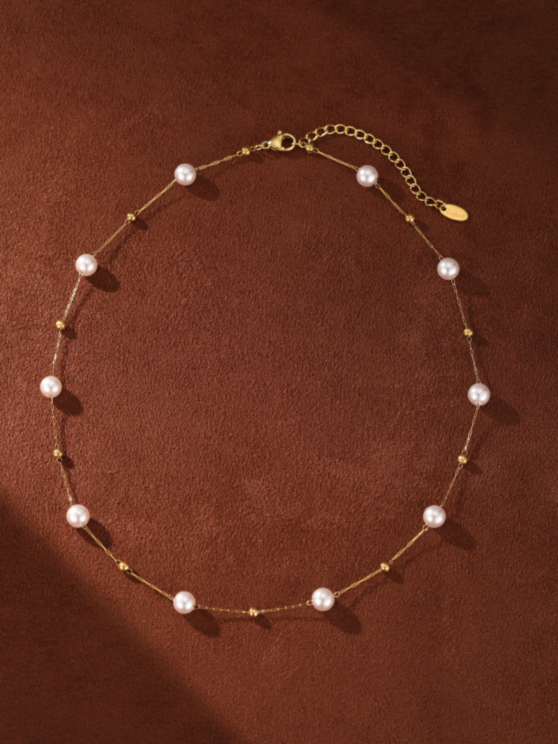 Un délicat collier à chaîne ton or présentant de petites perles rondes d'eau douce et de minuscules perles dorées, disposées comme si elles flottaient le long de la chaîne. Le collier est présenté sur fond marron chaleureux et texturé avec de douces ombres.