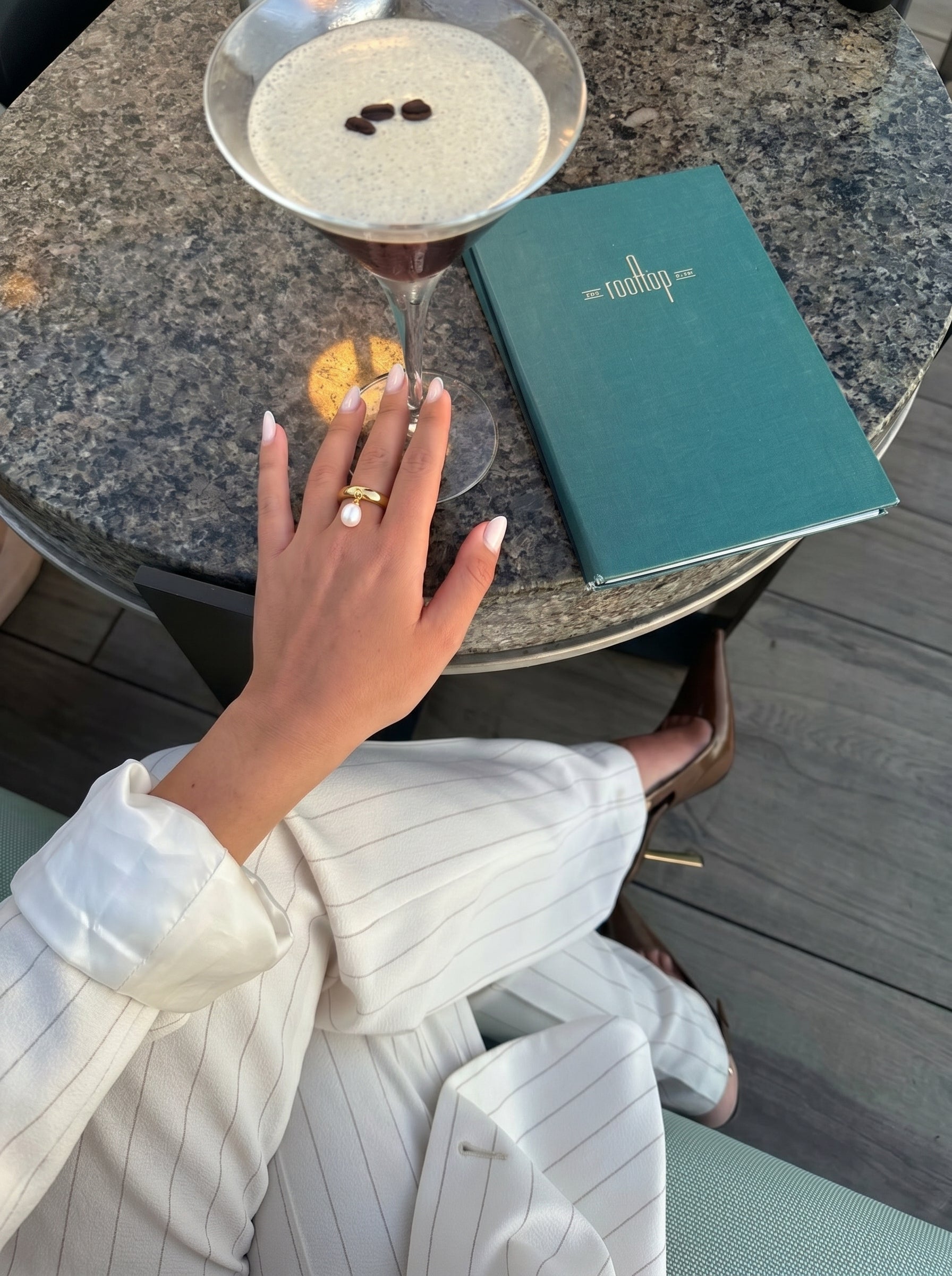 Close-up of a woman's hand wearing a gold ring with a dangling freshwater pearl, with a cocktail and a menu on a table. @lifestyle #lifestyle