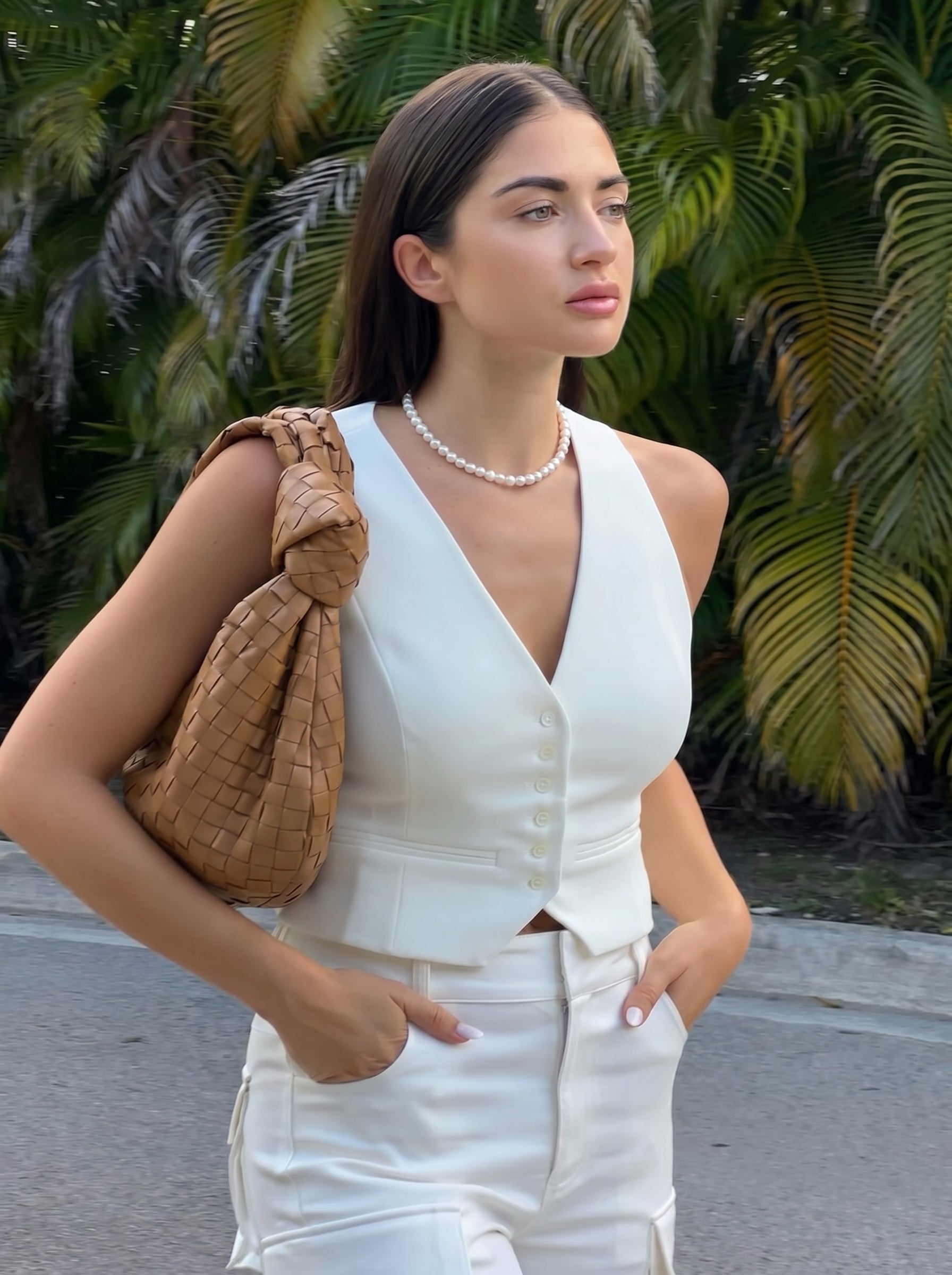 Une femme portant un gilet et un pantalon blancs, avec un collier ras-de-cou en perle d'eau douce et un sac à main tissé beige, debout à l'extérieur avec des palmiers en arrière-plan. @lifestyle